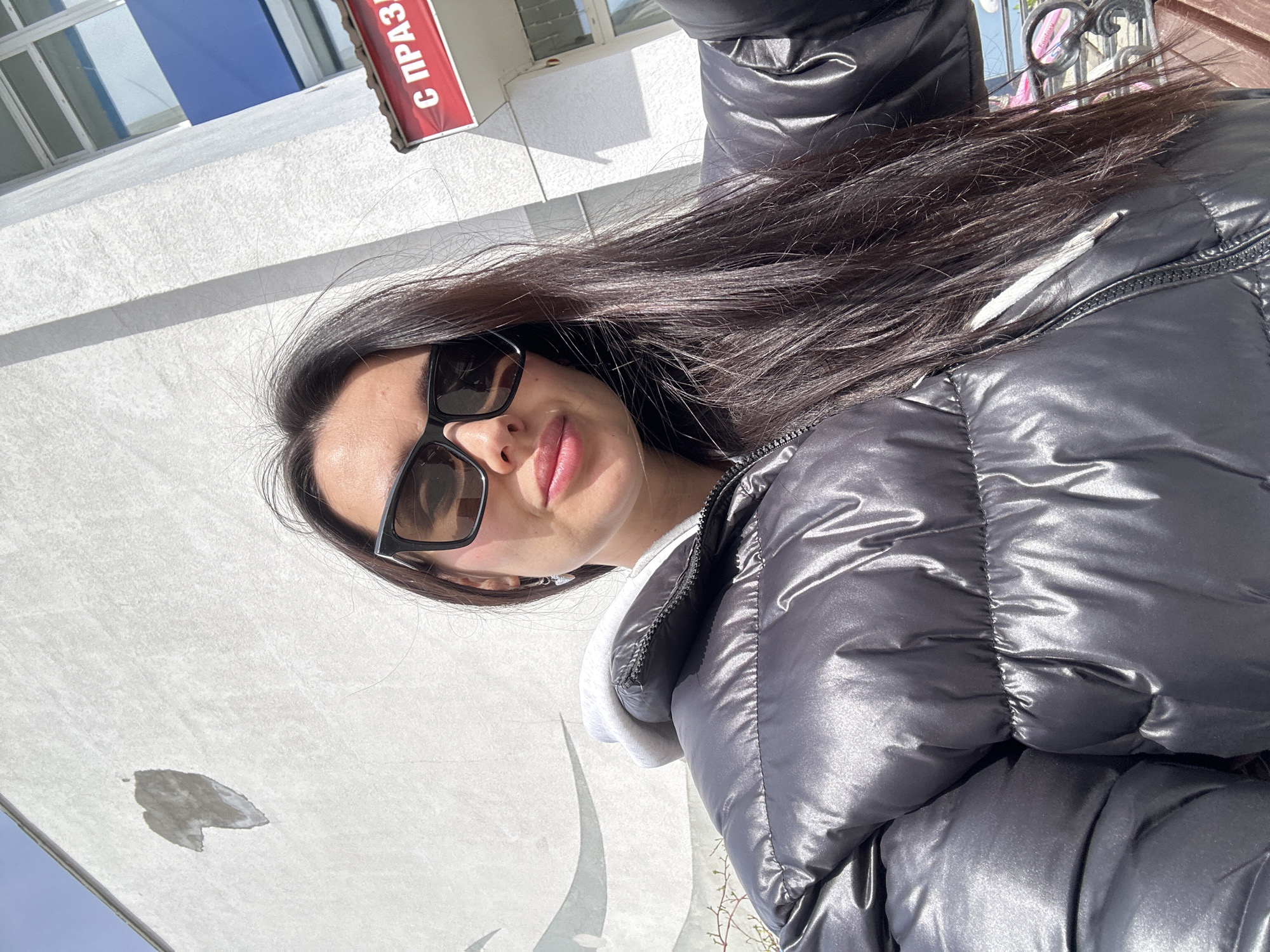 An outdoor selfie captured in Chaikovsky, Russia, features a young woman centered in the frame. She has long, dark hair flowing over her shoulders and is wearing large, dark rectangular sunglasses. Her lips appear to have a glossy finish. She is dressed in a shiny black puffer jacket, which reflects the bright sunlight, layered over a light-colored (possibly white or light grey) hooded sweatshirt, visible around her neck and part of her sleeve.
The background consists primarily of a plain white building wall, which is brightly lit by the sun, creating distinct shadows. On the upper left side of the image (when oriented correctly), a window with a blue panel is visible on the building. To the right of that, a red banner is attached to the building, displaying partially visible Cyrillic text: "С ПРАЗДН", which translates to "Happy Holiday!" or "Congratulations!". Below the woman, on the building wall, there's an irregular dark stain or patch. Sparse, twiggy branches of a plant are visible in the lower right corner, indicating some natural elements in the urban setting. The strong shadows and bright light suggest it is daytime, likely a sunny day. The woman appears to be posing for the camera, her expression neutral or slightly smiling.
