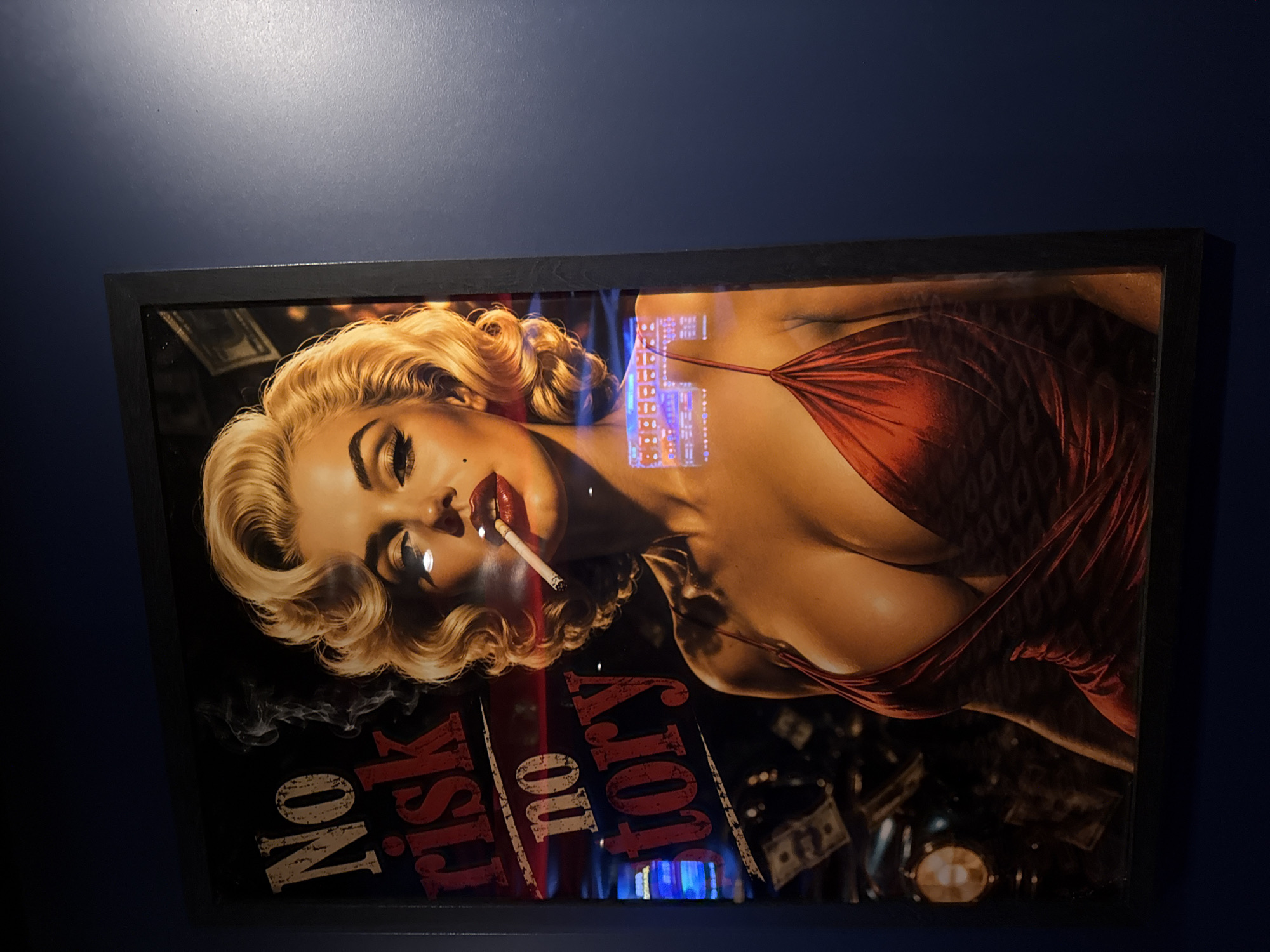 A framed print of Marilyn Monroe smoking is hung on a dark blue wall. The artwork has a black frame. The print features a blonde woman, resembling Marilyn Monroe, in a red dress, lying down and smoking a cigarette. The print also features the words "No Risk, No Story". The image includes some light reflections. The location is likely Burgas, Bulgaria, based on previous context.