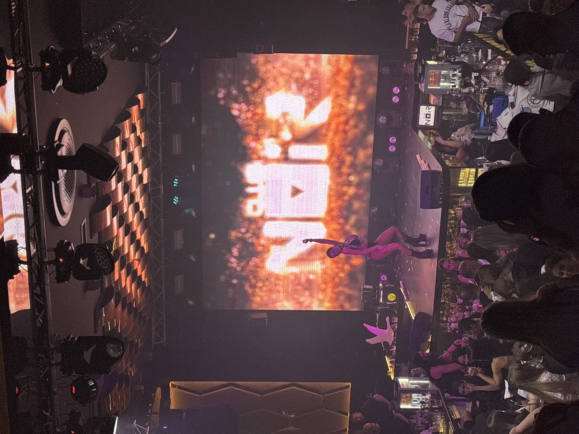The image depicts an indoor scene at a nightclub, possibly named "Club Noir" as indicated by signage on a screen and a bar counter. The setting appears to be lively, with a stage featuring a performer in a pink outfit in front of a large screen displaying bright, abstract graphics. There is a bar area to the left and in the foreground, with patrons seated and standing around tables, some of whom are looking towards the stage. The lighting is dim, with spotlights and colorful stage lights illuminating the area. The overall atmosphere suggests a nighttime entertainment venue. The city of Kazanlak, Bulgaria is not depicted or suggested in the provided image.