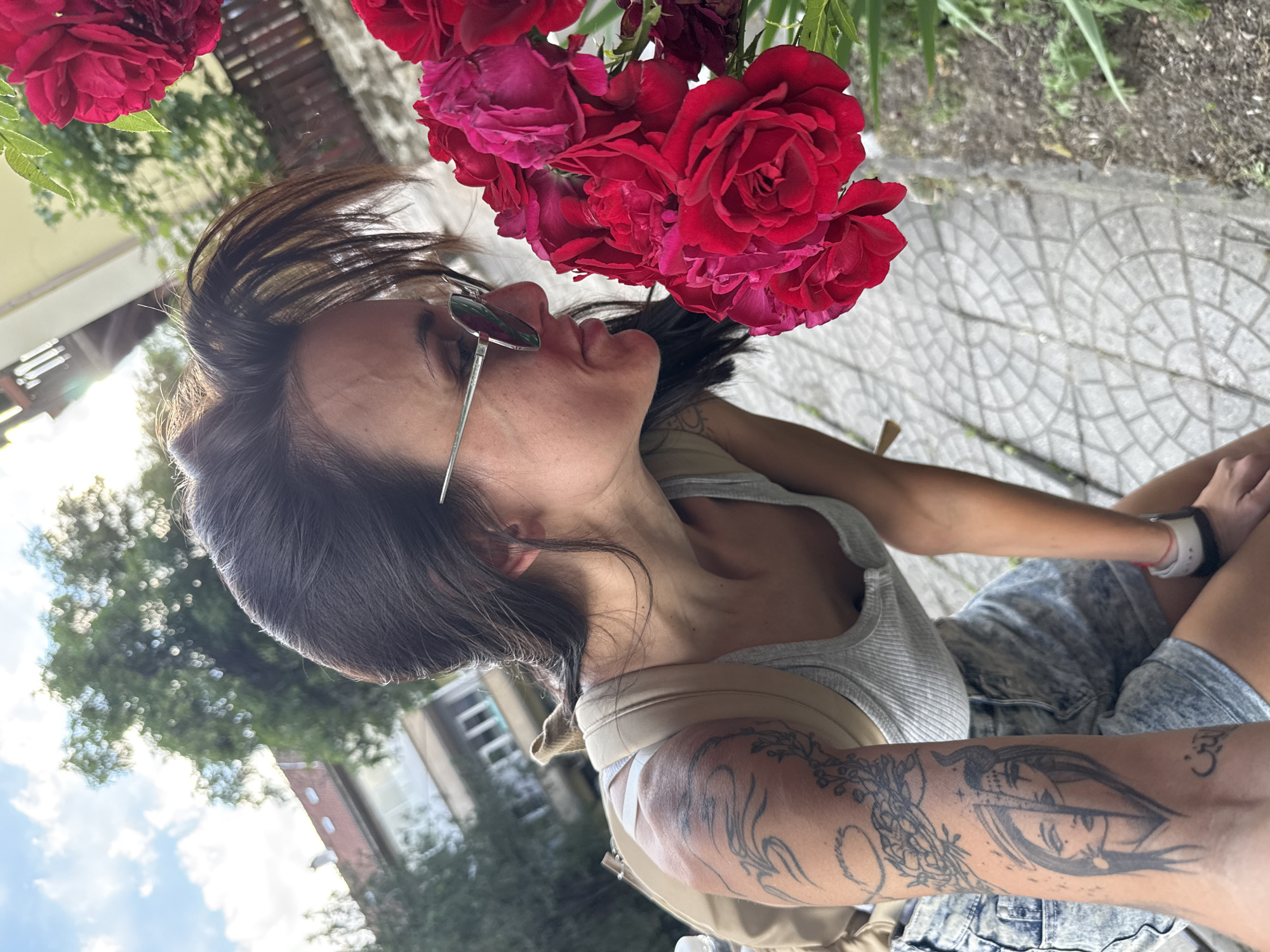 A woman is captured in profile, bending forward to smell a cluster of deep red roses. She wears a light grey ribbed tank top and light-wash denim shorts. A beige backpack strap is visible over her left shoulder. Her dark hair is pulled back, with loose strands framing her face and forehead. Mirrored aviator-style sunglasses are positioned on her nose, obscuring her upper eyes.
Prominent tattoos are visible on her left arm. On her upper arm, there are abstract and floral designs. On her forearm, a detailed tattoo depicts a split female face, one half appearing serene and the other with a subtle horn, with Arabic script visible beneath it. Her mouth is slightly open, suggesting she is inhaling the scent of the flowers.
The roses, positioned in the lower right foreground, consist of several vibrant red blooms. The scene is set outdoors on a bright day in an urban environment in Pleven, Bulgaria. The ground beneath her consists of grey paving stones with an interlocking hexagonal or circular pattern, bordered by a narrow strip of dry earth and sparse vegetation to the right.
In the background, out-of-focus green foliage from trees is visible, with noticeable sun flare indicating strong daylight. Behind the trees and to the upper left, an older multi-story building with a red-tiled roof is partially visible. Further to the right, a darker building and a wooden fence are blurred. The sky in the upper left displays patches of blue with white clouds.