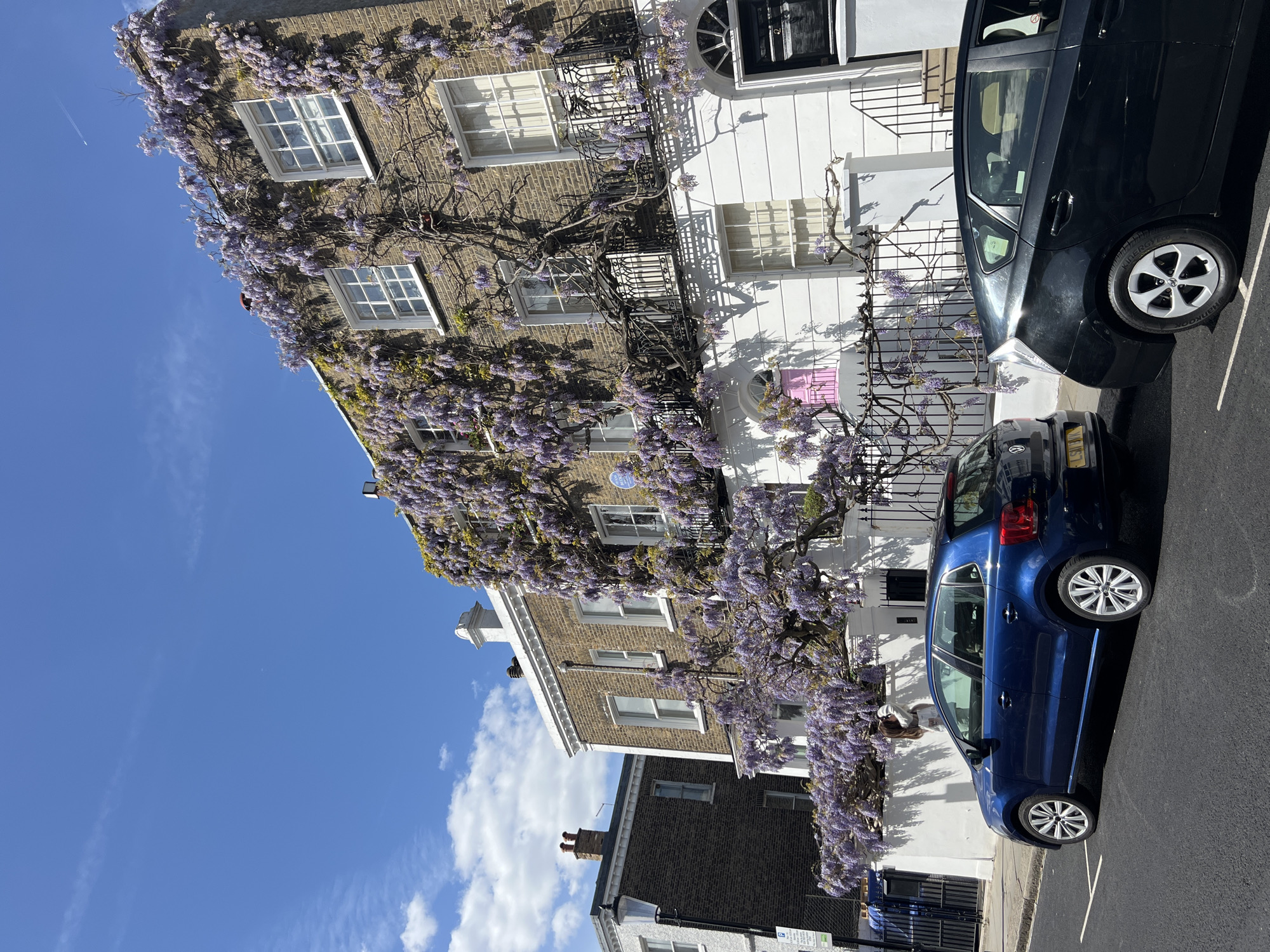 A street-level, eye-level, outdoor medium shot shows two buildings and two cars in the city of London, United Kingdom. The two buildings are on the left of the image. The buildings are two and three stories high. The higher building is made of brick and covered with wisteria. The lower building is black and white with a small window. Two cars, one blue and one black, are parked on the right of the image. The sky is blue with thin white clouds.