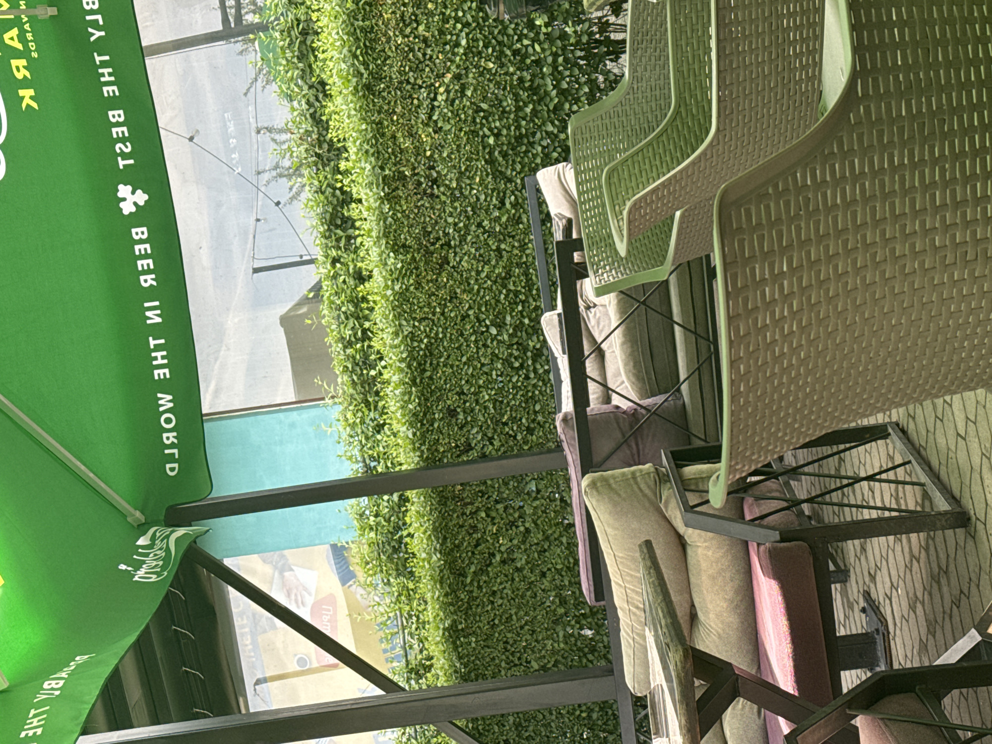 This media file captures an outdoor seating area, likely a cafe or restaurant patio, situated in Sofia, Bulgaria, during bright daylight. The scene is devoid of people, presenting a static view of the unoccupied space.
In the left foreground, a large, vibrant green umbrella provides shade. Text is visible on its surface, appearing mirrored due to the image's orientation. Phrases readable include "MARK," "THE BEST JAY," and "BEER IN THE WORLD," alongside a white clover-like symbol. Additional blurry mirrored text is also present further down the umbrella's canopy.
To the right, several patio chairs made of a green woven material are arranged around black metal tables. Many chairs are fitted with cushions in shades of beige, grey, purple, and a pinkish hue, indicating a comfortable setting. The ground beneath the seating area is paved with textured, possibly hexagonal or irregular, tiles.
A tall, dense hedge of small-leaved green foliage forms a natural backdrop, separating the patio from the background. Beyond the hedge, parts of a building or structure are visible, along with a blurry sign or poster that contains some Cyrillic text, potentially including words like "Пътят" (The Way) and "Плъм" (Plum). The overall impression is an inviting and ready-to-use outdoor space.