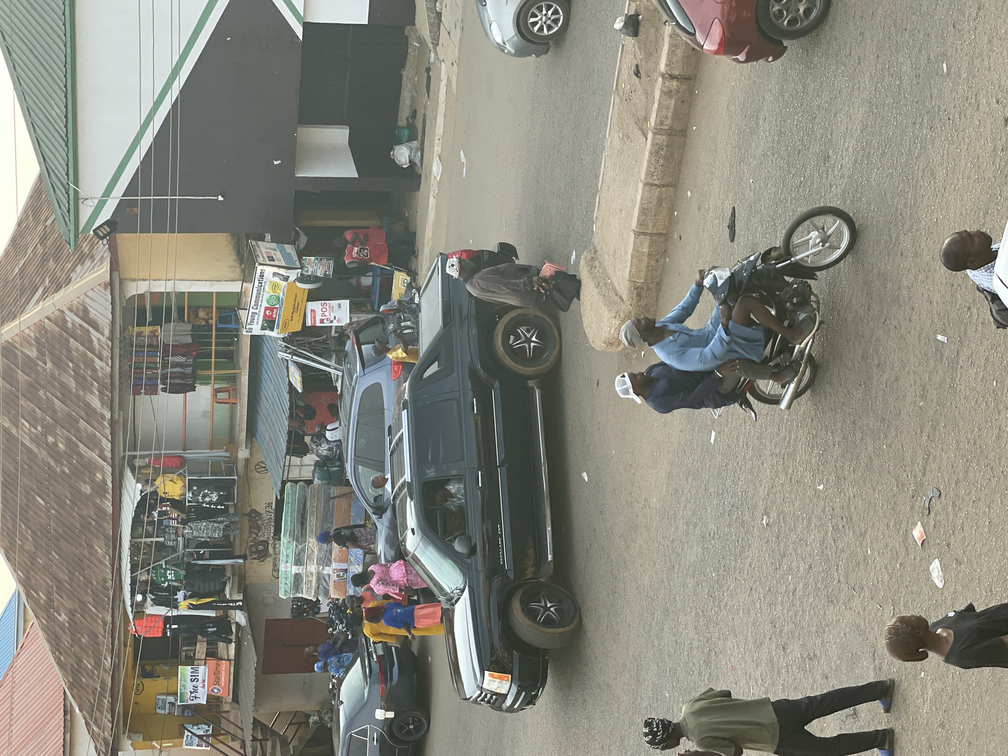 The image captures a street scene in Bukuru, Nigeria. The street is lined with storefronts selling goods like clothing and mattresses. Cars and a black Cadillac Escalade EXT are parked along the side of the road. A man is riding a motorcycle, and several people are walking along the street.