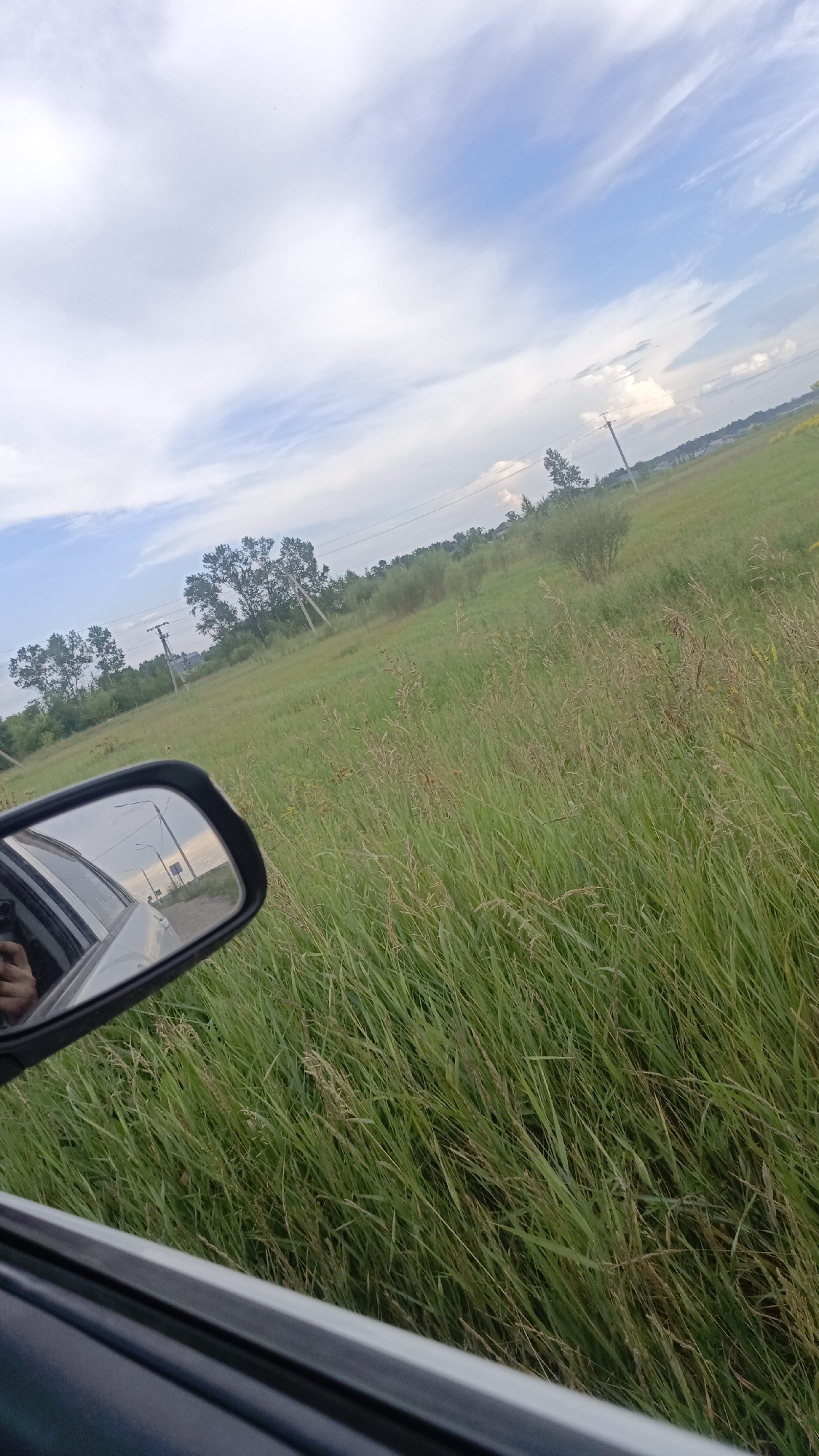 The image displays a view from inside a vehicle, looking out towards a rural landscape. In the foreground, a side-view mirror reflects a hand. The mirror also shows a road with utility poles and a street light. Beyond the vehicle, a vast field of tall, green grass stretches towards a line of trees in the middle ground. The background consists of a blue sky with scattered white clouds. The overall scene suggests a drive through a country or suburban area, possibly during the daytime. There are no visible landmarks to identify the specific location as Angarsk, Russia.