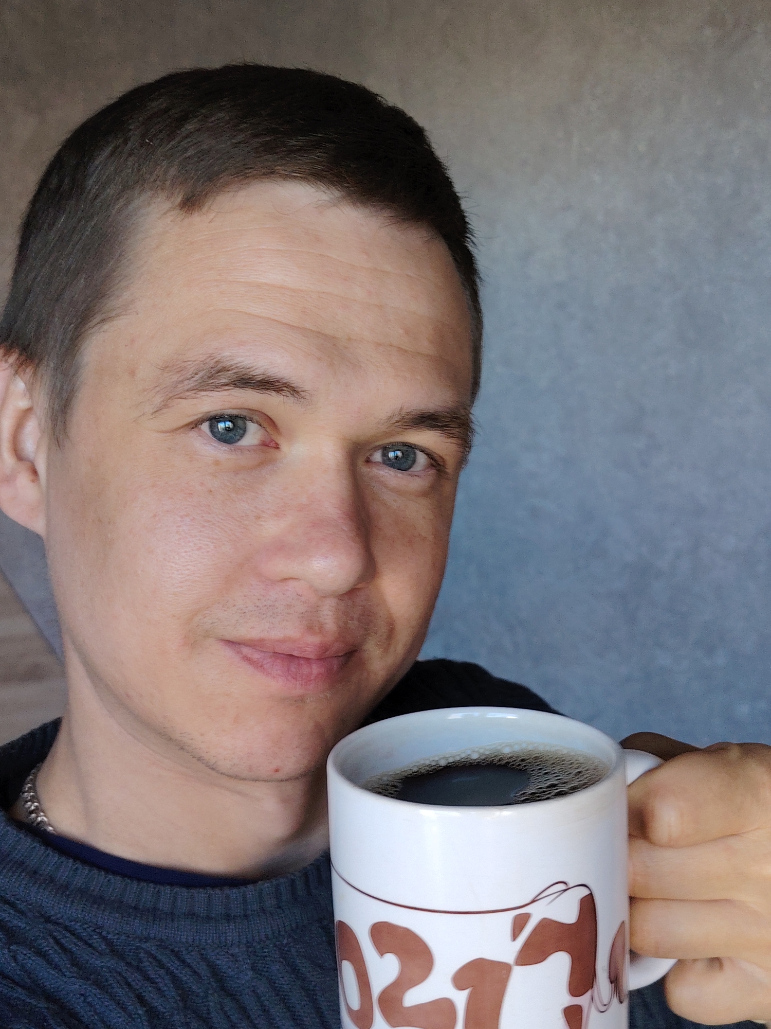 This close-up, half-portrait captures a light-skinned man holding a mug of coffee indoors. The man, appearing to be in his late 20s or 30s, has short, dark brown hair and striking blue eyes, looking directly at the viewer with a gentle, faint smile. He sports faint stubble and natural skin texture.
He is dressed in a dark grey, possibly knitted, sweater over a dark blue collared shirt, with a silver chain visible around his neck. In his right hand, he holds a white ceramic mug filled with dark coffee, topped with a thin layer of foam. The mug features prominent brown designs, clearly displaying the number "21" alongside other abstract brown shapes that resemble cow spots or a stylized cow's head or ears.
The background is a simple, textured light grey wall, typical of an indoor setting. The overall impression is one of calm contentment, suggesting a casual moment, perhaps a coffee break during the daytime in Chaikovsky, Russia. No specific time of day or outdoor weather cues are visible.