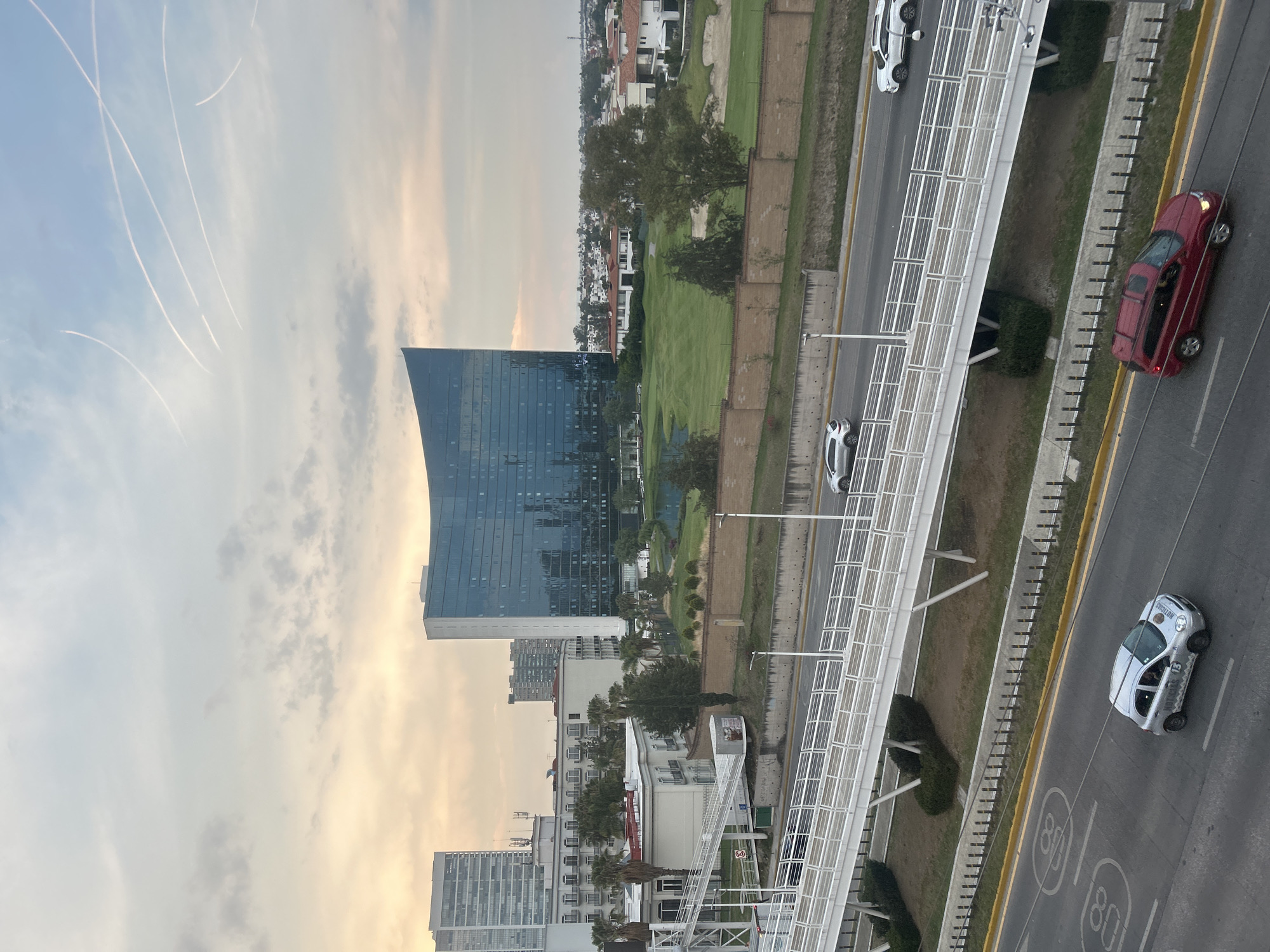An aerial view captures an urban and suburban landscape under a late afternoon or early evening sky. The sky, occupying the upper portion of the frame, is a soft blue with scattered light clouds and numerous white contrails crisscrossing.
In the midground, a prominent modern skyscraper with a curved glass facade stands on the left. To its right, a large green space, possibly a golf course or park, is visible, bordered by residential houses and smaller buildings further in the background.
The foreground is dominated by a multi-lane highway running horizontally across the image. A white pedestrian bridge, featuring a distinctive lattice-like structure, spans over the highway. Several cars are present on the road: a red sedan with its brake lights illuminated, a silver hatchback, and a white sedan. The silver hatchback has the word "PRECISION" written on its side. Large white markings of "80" are painted on the road, indicating the speed limit.
The scene depicts a blend of commercial and residential areas, suggesting a developed city with well-maintained infrastructure and green spaces. The overall impression is calm, with no specific activity beyond the movement of vehicles implied by their presence on the highway.