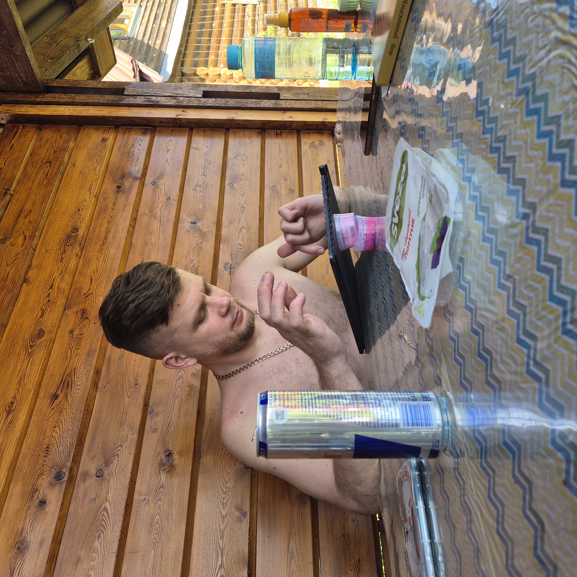 An adult male is seated shirtless at a table within a wooden structure in Новостройка, Russia. He has short, dark brown hair, a light beard, and a gold-colored chain around his neck. His right shoulder exhibits faint reddish marks. He is leaning forward, interacting with a black tablet computer placed on the table, utilizing both hands. The tablet is propped up by two stacked transparent pink plastic containers.
The setting is a semi-enclosed space constructed from horizontally aligned wooden planks, stained in a warm brown tone with visible grain and knots. To the right, an opening reveals exterior wooden construction, a green roof, and a distant window.
The table is covered with a reflective, transparent material over a patterned tablecloth featuring blue, yellow, and gray chevron designs. Items on the table include:
* To the left, an opened silver Red Bull energy drink can with Cyrillic text and a barcode-like pattern, next to which a smartphone in a clear case with a cartoon design is partially visible.
* In the center, a white and green flexible pouch with Cyrillic text reading "Svea" and "Прованские травы" (Provence herbs).
* To the right, a clear plastic water bottle with a blue cap and Cyrillic label, an orange spray bottle, a small green circular container, and a light yellow book with Cyrillic text "ИЗЫСКАННЫЙ ВАШ" (Your Exquisite) reflecting light. A dark-colored smartphone lies flat on the table, reflecting the surroundings.
The scene is illuminated by natural light, casting soft shadows. Reflections of various objects and the male's torso are visible on the table's surface.