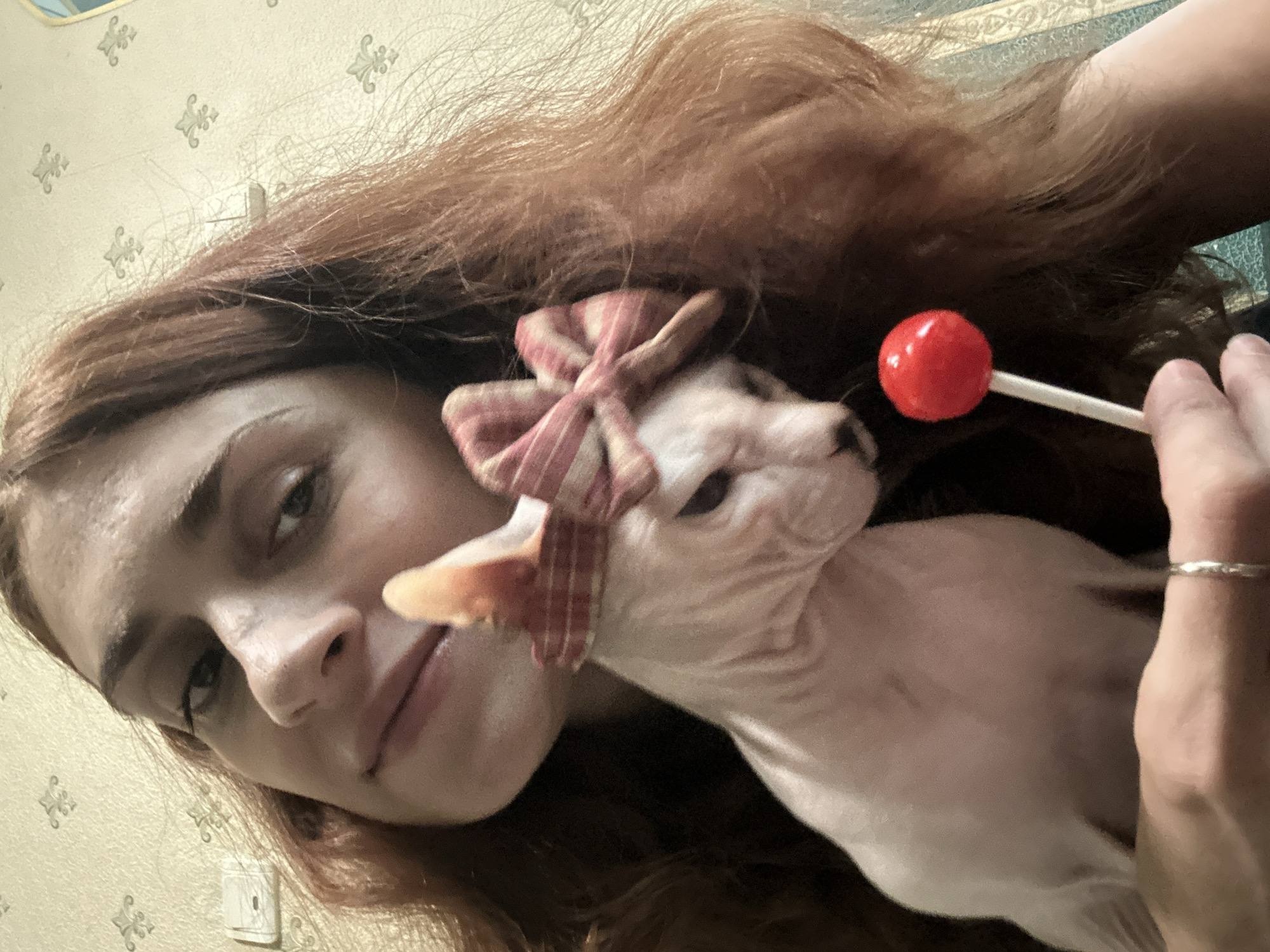 A woman with long, auburn hair is taking a selfie. She is holding a hairless cat, which is wearing a pink and white striped bow on its head. The cat's head is tilted down, and it appears to be looking at a red lollipop that the woman is holding up to it. The woman is smiling slightly. The background appears to be a wall with a patterned wallpaper. The image was taken indoors. There is no information in the image to suggest it is in Togliatti, Russia.
