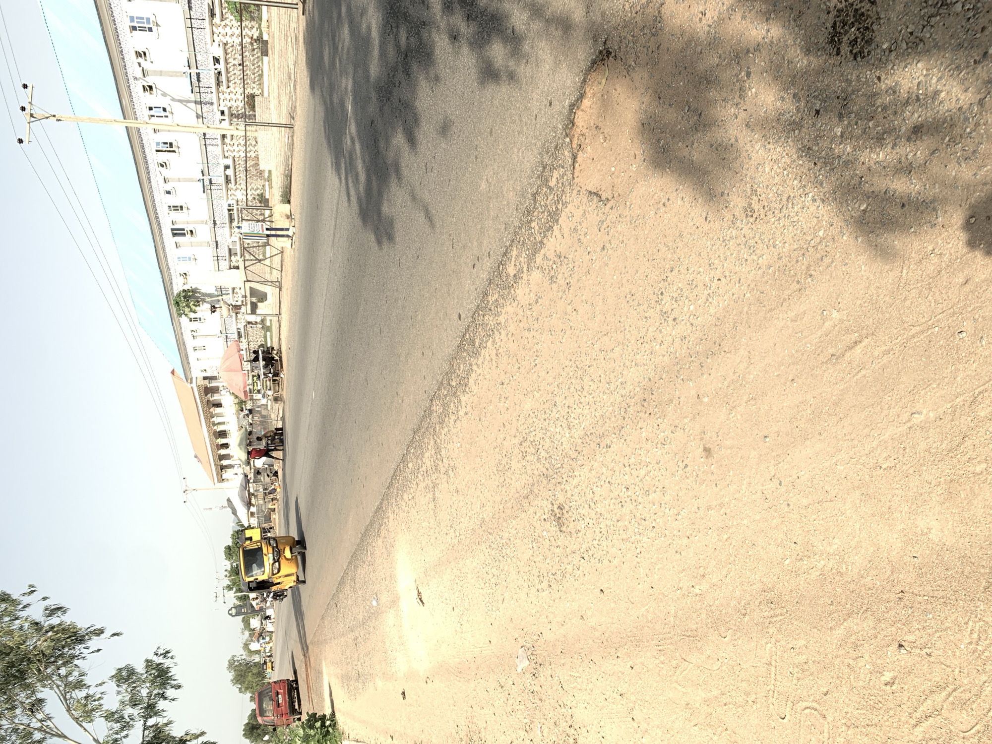 The image depicts a street scene in Jalingo, Nigeria. A yellow tricycle, commonly known as an "Abajaj," is the central subject, moving along the paved road. To the left of the tricycle, a red van is parked. On the right side of the road, a long, white building with a light blue roof stretches into the distance, featuring numerous windows and architectural detailing. In front of the building, there is a fence and several small stalls with umbrellas, suggesting a marketplace or roadside vendor area. A few individuals are visible near these stalls. The road is bordered by a sandy, unpaved shoulder on the left. The sky is clear and bright, indicating a sunny day. The overall environment appears to be an urban or semi-urban setting within Jalingo.