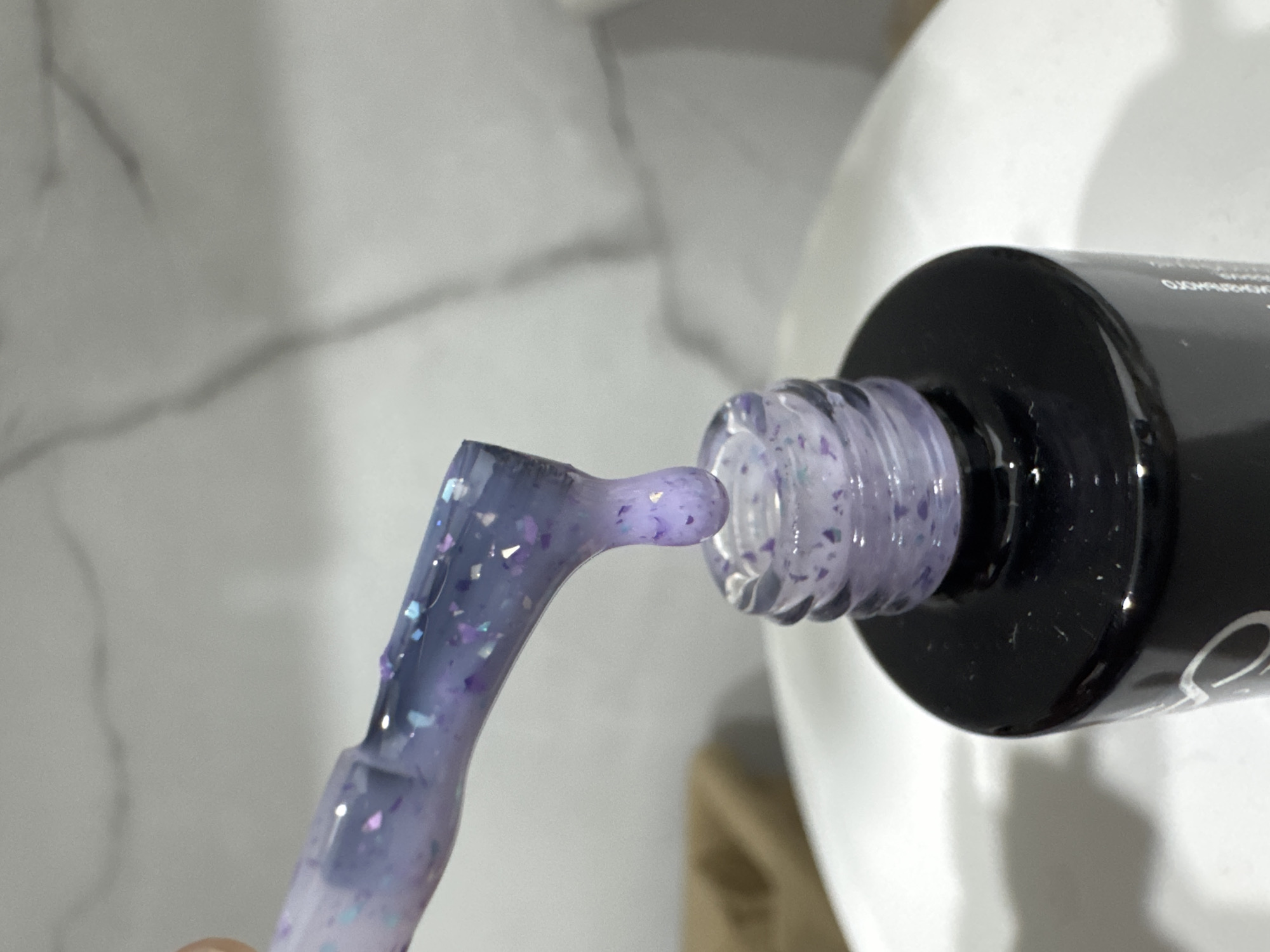 The image shows a close-up of a nail polish bottle with a brush applicator. The nail polish is a translucent purple with flecks of glitter. The brush is coated in the polish and is positioned near the opening of the bottle. The background is a blurred marble-like surface. The lighting suggests an indoor setting. There is no visible text in the image. The image focuses on the detail of the nail polish and its application tool. The scene is static, depicting a moment of preparation or use of the product.