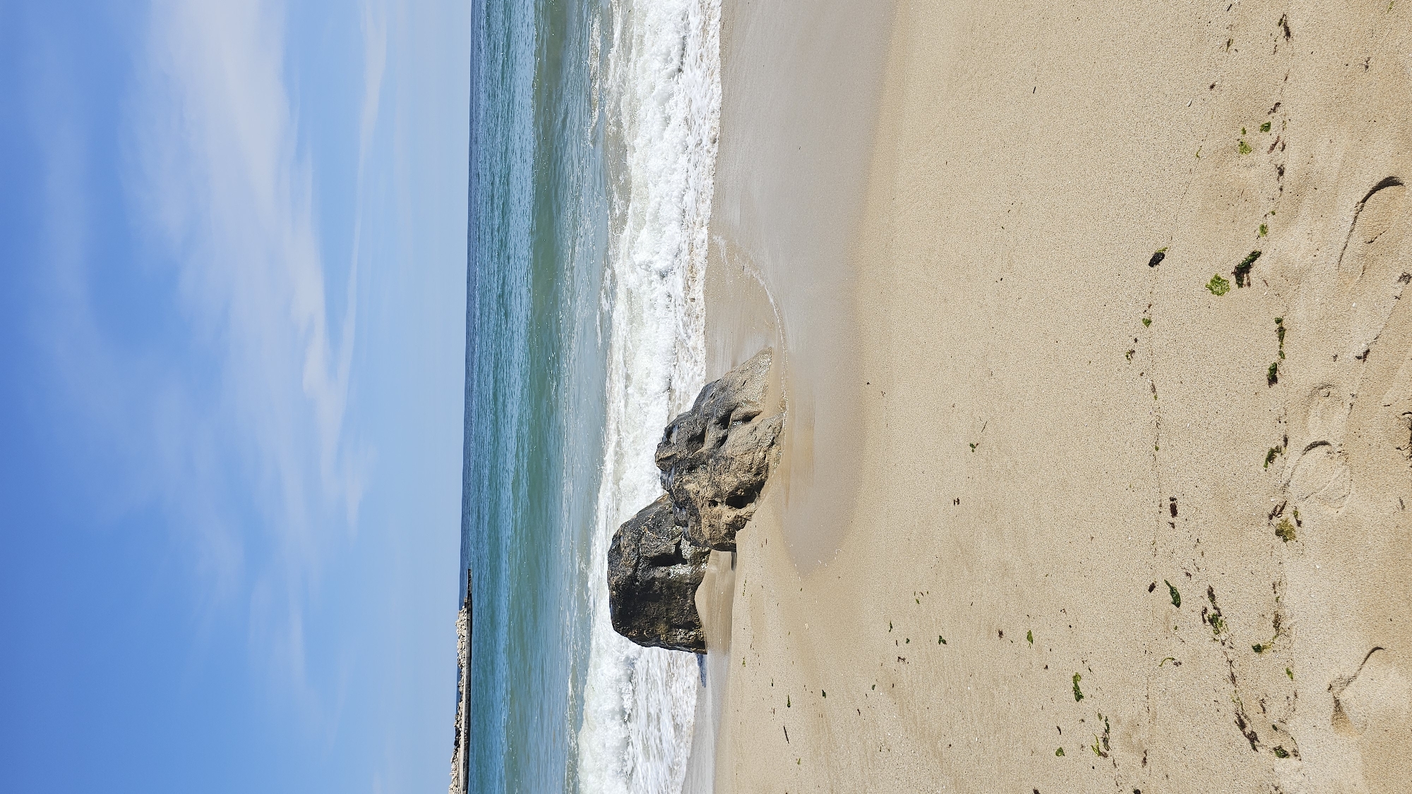 A tranquil, horizontal landscape photograph captures a peaceful beach scene in Varna, Bulgaria, under a clear sky. The image is divided into three primary horizontal elements: the sky, the sea, and the sand.
The upper portion of the frame features a bright, azure blue sky with wispy white clouds scattered across it, indicating sunny and pleasant weather conditions.
Below the sky, the calm sea displays a gradient of colors, moving from a deeper blue further offshore to a lighter, clear turquoise closer to the beach. Gentle waves lap against the shore, forming white foam where they meet the sand. On the far left, a low-lying breakwater or pier structure extends into the water, hinting at a coastal development.
The most prominent section of the image is the wide expanse of light, golden-beige sand. Two large, dark grey rocks are positioned near the waterline, partially submerged by the retreating waves, their surfaces glistening with moisture. Numerous distinct footprints are visible in the damp sand, predominantly on the right side of the frame, suggesting someone recently walked along the shore. Small clumps of green seaweed or algae are scattered across the sand, carried in by the tide.
No people are present in the photograph, conveying a sense of serene solitude. The scene depicts a bright, daytime setting, emphasizing the natural beauty and calm atmosphere of the Bulgarian coast. There is no visible text in the image.