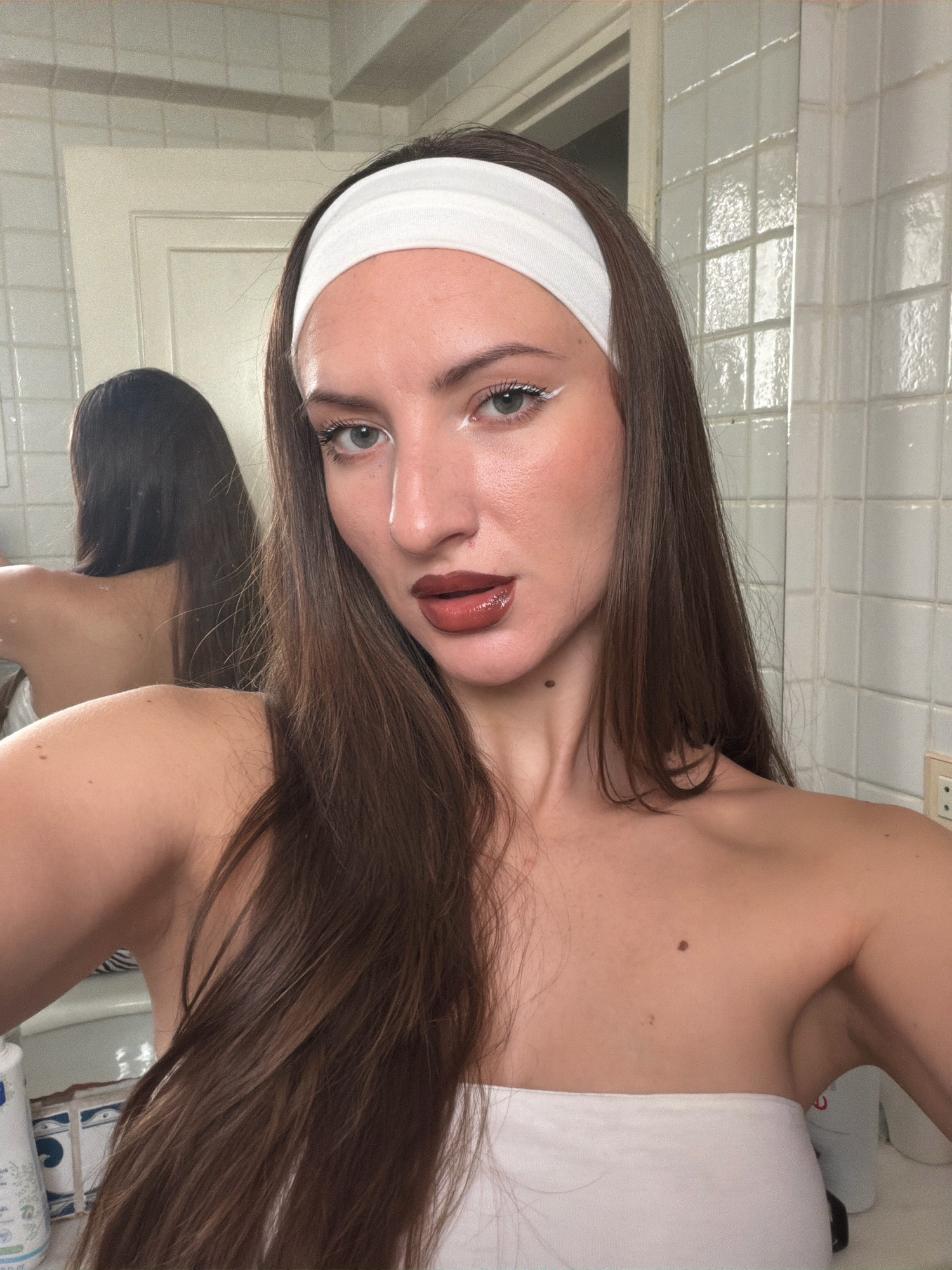 A woman with long, dark brown hair and a white headband is looking at the camera, her face partially obscured. She is wearing dark lipstick and white eyeliner. Her shoulders are bare, and she is wearing a white bandeau top. The background appears to be a bathroom with white tiled walls and a mirror reflecting her back. The image is taken at close range, focusing on her face and upper body. There is no indication of the city of Sharm El Sheikh, Egypt, within the image.