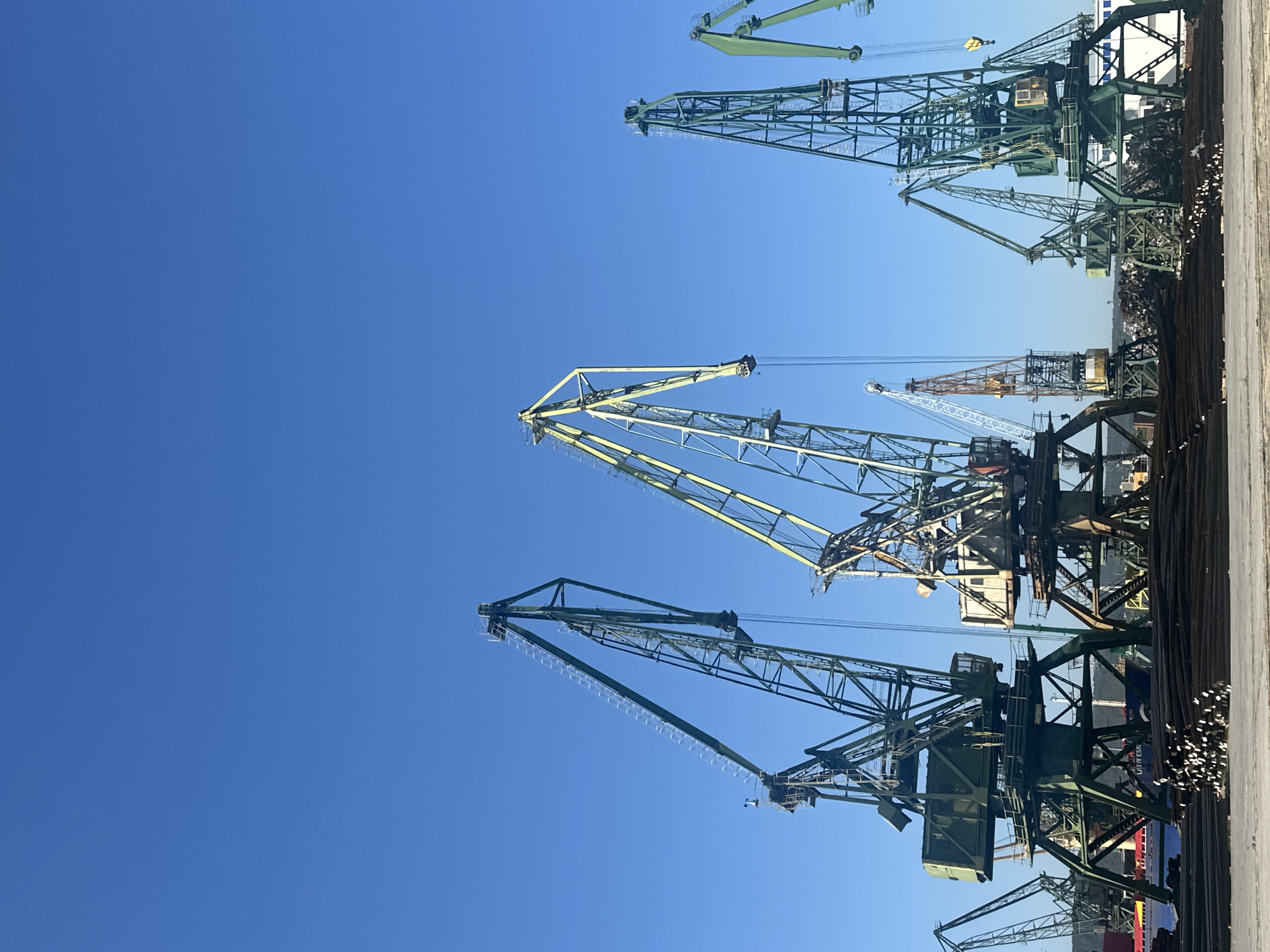 A wide-angle, low-angle shot captures a clear, bright blue sky dominating the upper two-thirds of the frame. Below the sky, several industrial port cranes are visible, their metallic structures silhouetted against the sky. The cranes are of varying sizes and designs, with some being predominantly dark green and others featuring yellow accents. They stand on sturdy bases and are interconnected with complex metal frameworks. In the foreground, a pile of dark metal rods or pipes is neatly arranged, with some sections showing white tips. In the background, a large ship with the word "VESK" visible on its hull is docked, and further back, a hazy line of land with hills is discernible. The overall scene depicts a busy industrial port with heavy machinery.