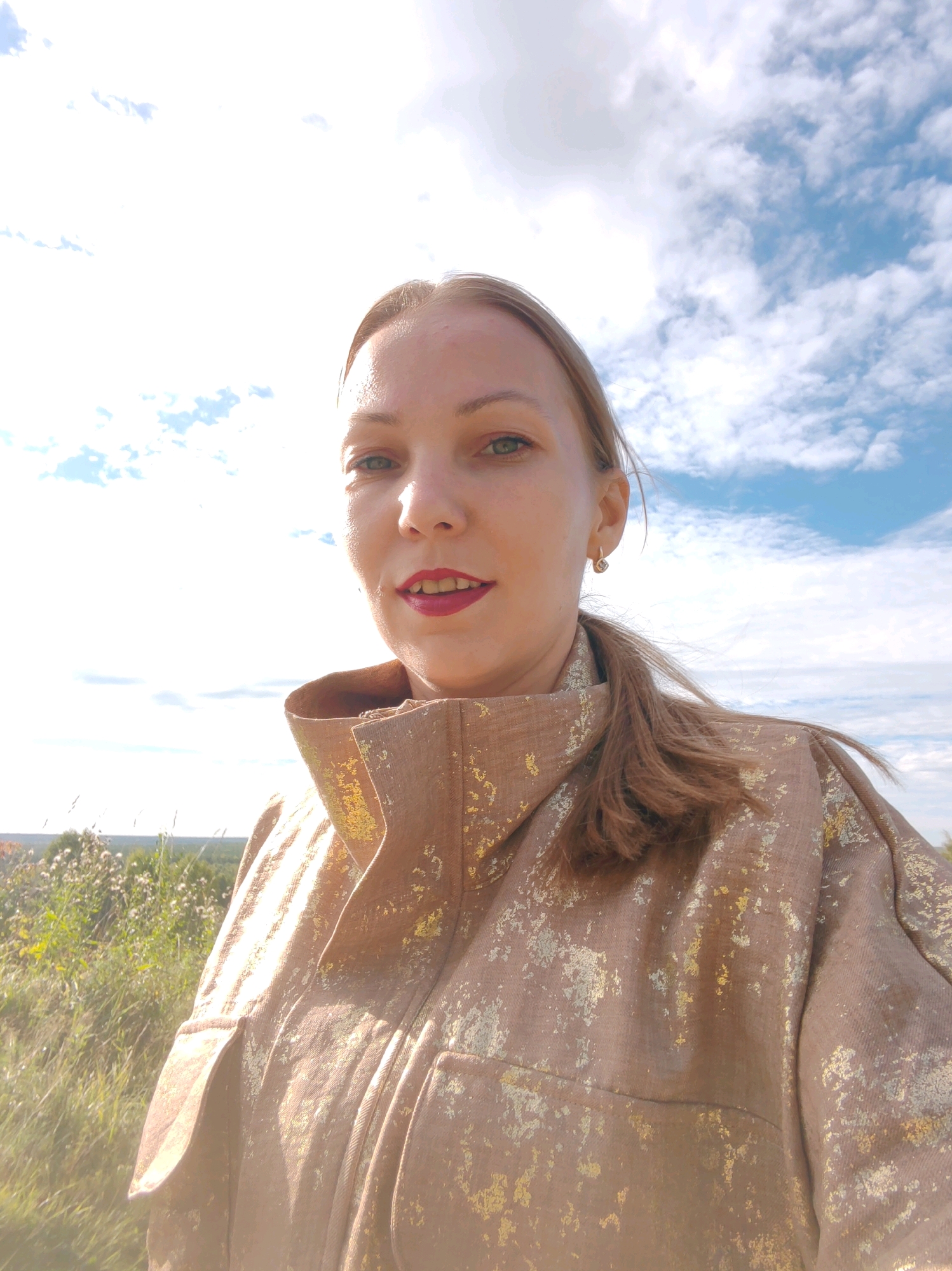 The image shows a close-up selfie of a Caucasian woman with blonde hair and green eyes. She is wearing a light brown jacket with a metallic gold pattern and red lipstick. The woman is outdoors, with a blue sky and white clouds in the background. In the foreground, tall grass and some wild flowers are visible. The scene appears to be a rural or natural setting, possibly a field or a hillside. There is no indication of it being in the city of Чухлэм, Russia.