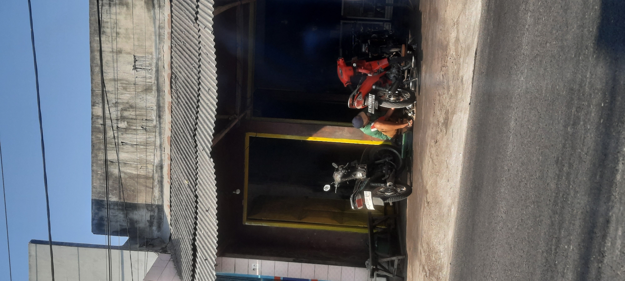 In the city of Selopuro, Indonesia, a man wearing a green shirt and brown shorts kneels on the ground in front of a red motorcycle, appearing to be working on it. He has a dark baseball cap on his head and is wearing sandals. To his left, a black motorcycle is parked. The motorcycles are positioned outside a building with a corrugated metal roof and a dark entrance with a yellow frame. A wooden bench is visible to the left of the entrance. The ground is paved with concrete, and a road is in the foreground. The scene is illuminated by natural daylight under a clear blue sky.