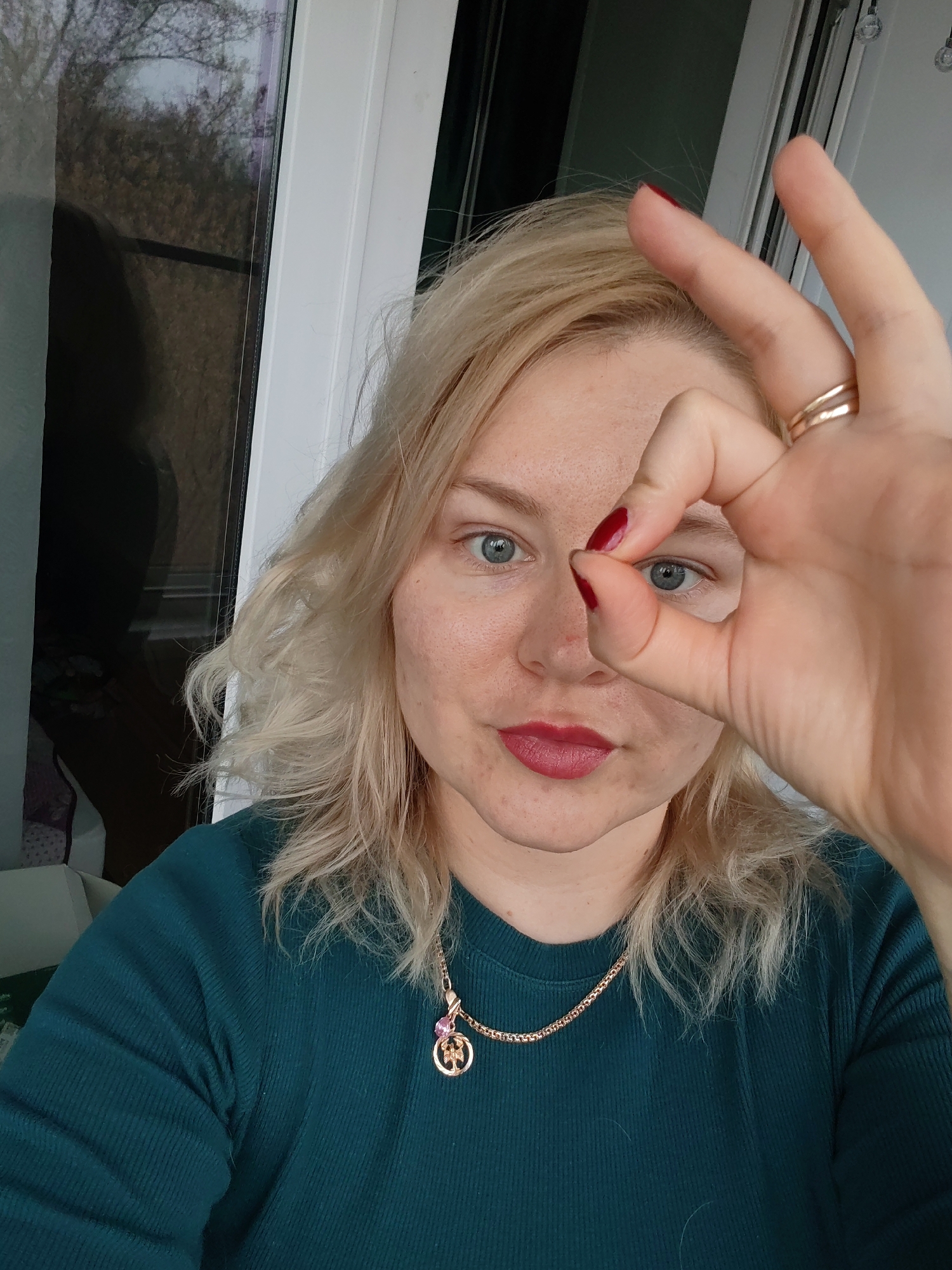 A Caucasian woman with blonde, shoulder-length wavy hair and blue eyes poses for a selfie. She is wearing a dark green ribbed top and a gold necklace with a round pendant. She has a gold ring on her index finger and red nail polish on her nails. She is making a circle with her index finger and thumb over her right eye. The background shows a white-framed window with a blurred outdoor scene. The location is inferred to be Izhevsk, Russia, based on the provided information but is not visually verifiable in the image.