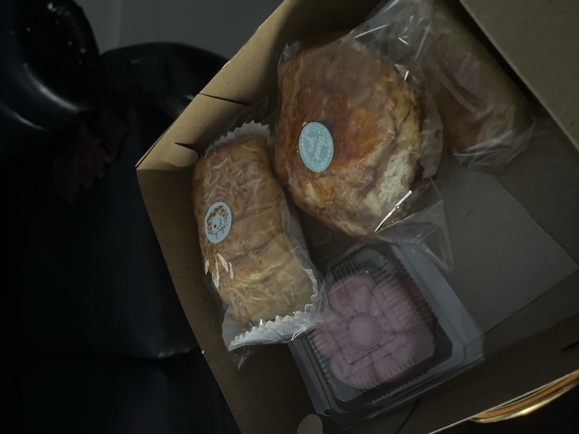 A cardboard box contains four baked goods. Two appear to be loaves of bread, one with a sprinkle of shredded cheese on top and a floral sticker that reads "Thank you". Another loaf has a similar sticker with floral elements. The third item is a square plastic container with pink, layered pastries. The fourth item is partially visible, also a baked good in plastic wrapping. The box is resting on a dark, possibly leather surface, and the background is out of focus, suggesting an indoor setting. The lighting is dim. There is no indication of the city of Jambearum, Indonesia, in the image.