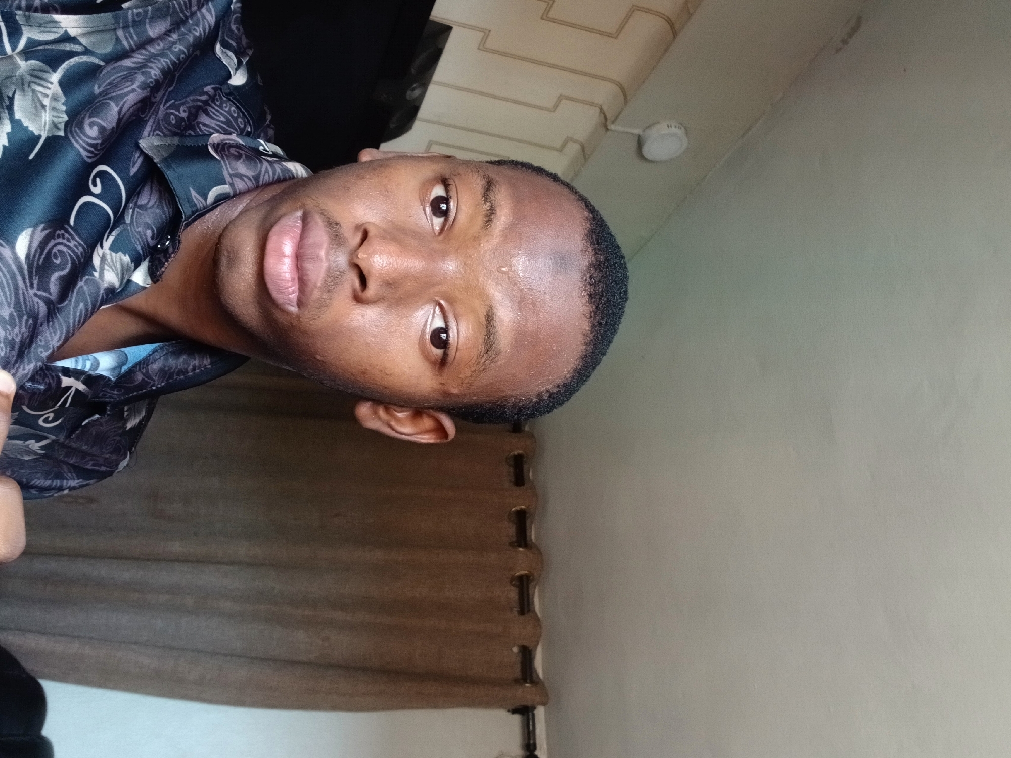 A dark-skinned man with a closely cropped hairstyle is photographed from a low angle indoors. He is wearing a dark patterned shirt with white floral designs. His face is moist, possibly with sweat, and he has dark brown eyes. The background includes off-white walls, a beige curtain or blind, and a portion of a ceiling with a light fixture. There is no verifiable evidence to confirm the location is Lagos, Nigeria.