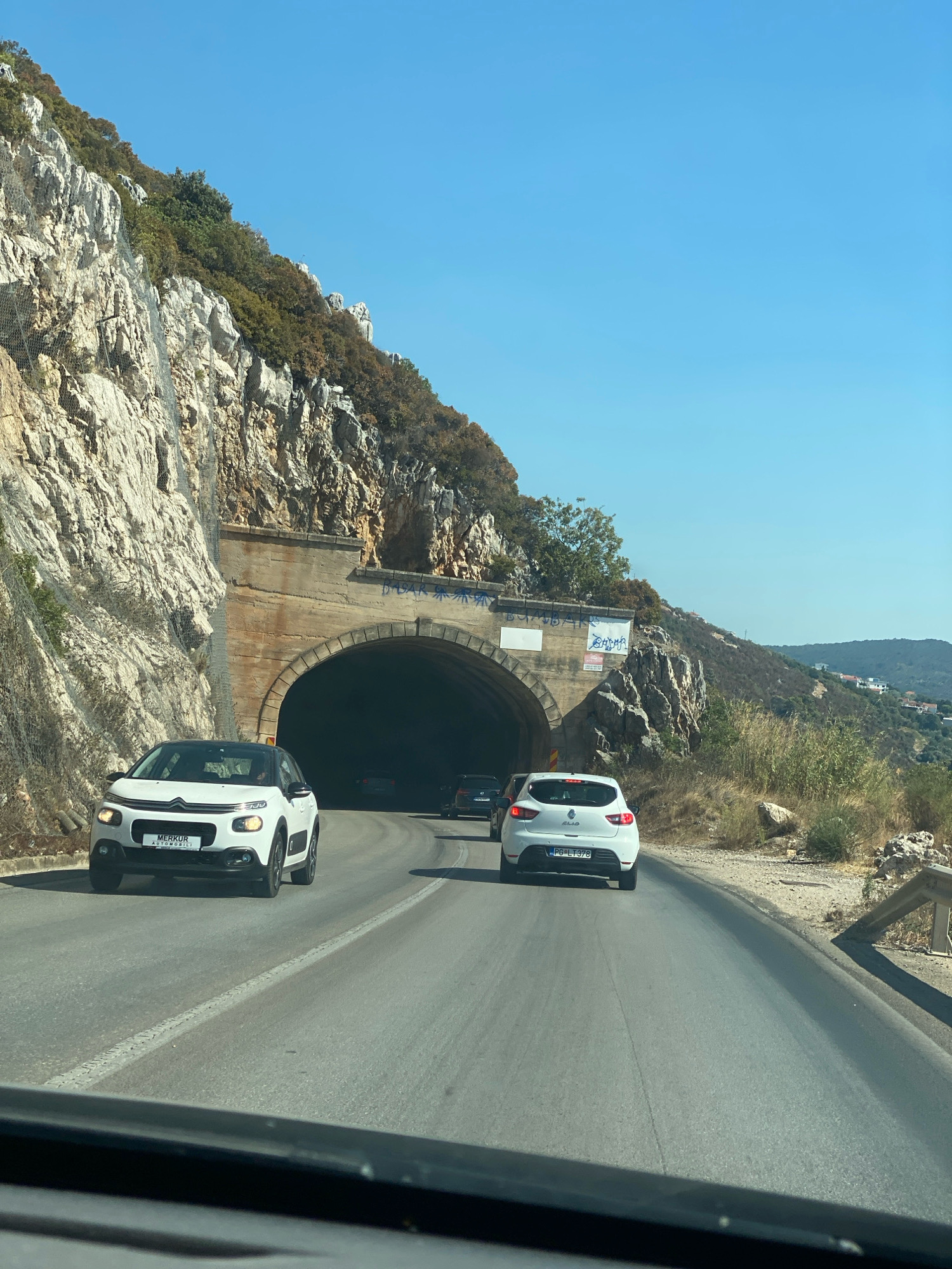 The image shows a sunny day with a clear blue sky. A road leads towards a tunnel carved into a rocky mountainside. The tunnel entrance is wide and arched, with concrete supports. A white Citroen C3 is driving on the road in front of the tunnel, and a white Renault Clio is behind it. Other cars are visible inside and approaching the tunnel. The mountainside is covered with sparse vegetation and rocks, and a metal safety net is installed on the cliff face. In the distance, hills with scattered buildings can be seen. The scene appears to be on a coastal road near Dobra Voda, Montenegro.