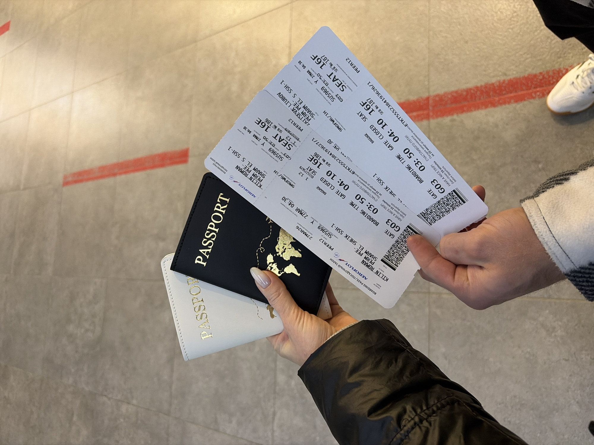 The image displays a close-up, downward-angled view of two individuals' hands holding travel documents within an airport terminal. The left hand, featuring manicured nails with light polish and partially covered by a black puffer jacket sleeve, securely holds two passport covers: one white and one black, both embossed with gold "PASSPORT" lettering and a gold world map graphic. This hand also holds the upper sections of two white paper boarding passes. The right hand, dressed in a grey and white checkered sleeve, grasps the lower portions of the same boarding passes.
The boarding passes, issued by Aeroflot, detail flights for "KILIN ROMAN" (seat 16E) and "ZAITSEVA LIUBOV" (seat 16F) on flight SU5969, scheduled for March 27, 2026. The flight originates from Perm, Russia, and is destined for Sharm El Sheikh, Egypt. Boarding time is 03:50, with the gate (G03) closing at 04:10 for a 04:30 departure. Each pass includes a QR code, flight specifics, and Russian text, notably "СЧАСТЛИВОГО ПУТИ!" (Have a nice trip!).
The floor beneath the hands is composed of light grey tiles with visible grout lines. Two parallel red lines are marked diagonally on the floor, suggesting a designated path or queue. A person's right foot, shod in a white sneaker with dark stripes, is partially visible in the bottom right corner of the frame. The scene captures the moment of preparing for an international flight departing from Perm, Russia, within a modern airport setting. The exact location in the city of Boldino, Russia, cannot be confirmed from the image; the flight originates from Perm.