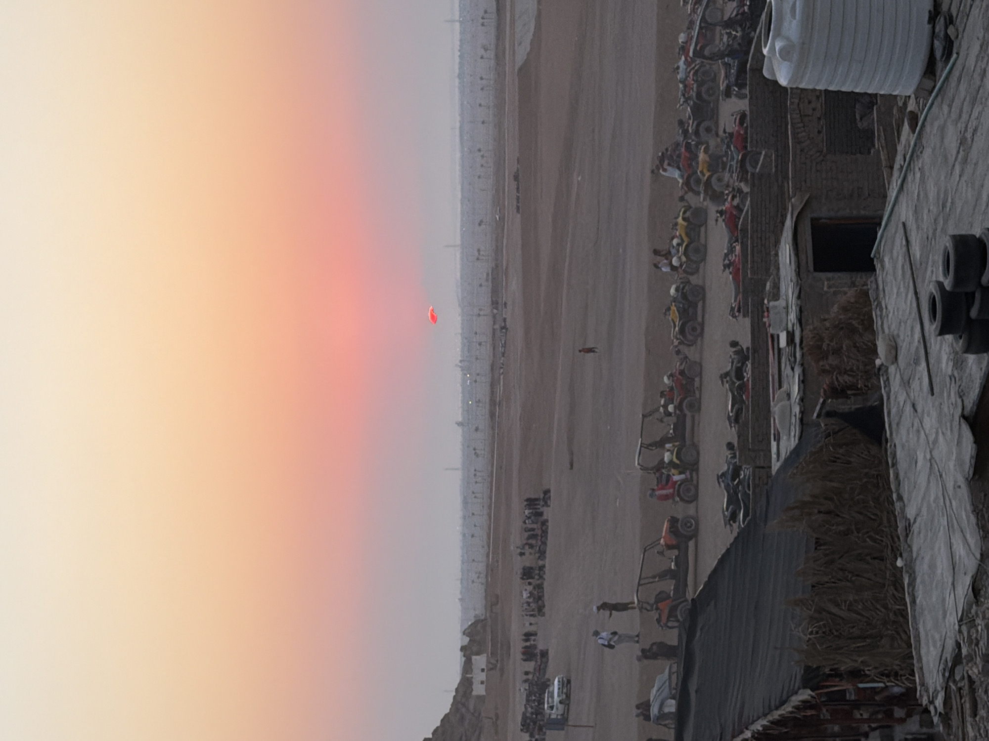 The image depicts a desert landscape at sunset, likely in Sharm El Sheikh, Egypt, characterized by its arid terrain and clear sky tinged with hues of orange and pink. In the foreground, a rustic, open-sided structure with a thatched roof is visible, constructed from natural materials. Adjacent to this structure, various ATVs and quad bikes are parked in neat rows, suggesting a recreational or tour operation. Several people are present in this area, some appearing to be staff and others participants, interacting with the vehicles and preparing for an activity.
Further in the middle ground, a larger gathering of people and ATVs is observable in a dusty, open area. A white pick-up truck is parked among them. In the background, a vast expanse of flat, sandy terrain stretches towards the horizon, dotted with a sparse distribution of utility poles or small trees. Beyond this, distant buildings and structures can be discerned, with a hazy outline of hills or mountains in the far distance.
The sky itself is a prominent feature, transitioning from a pale yellow at the top to vibrant oranges and pinks closer to the horizon, where the sun is setting. A small, bright red object, possibly a paraglider or a kite, is suspended in the sky. The overall atmosphere is one of an outdoor adventure venue at the end of the day.