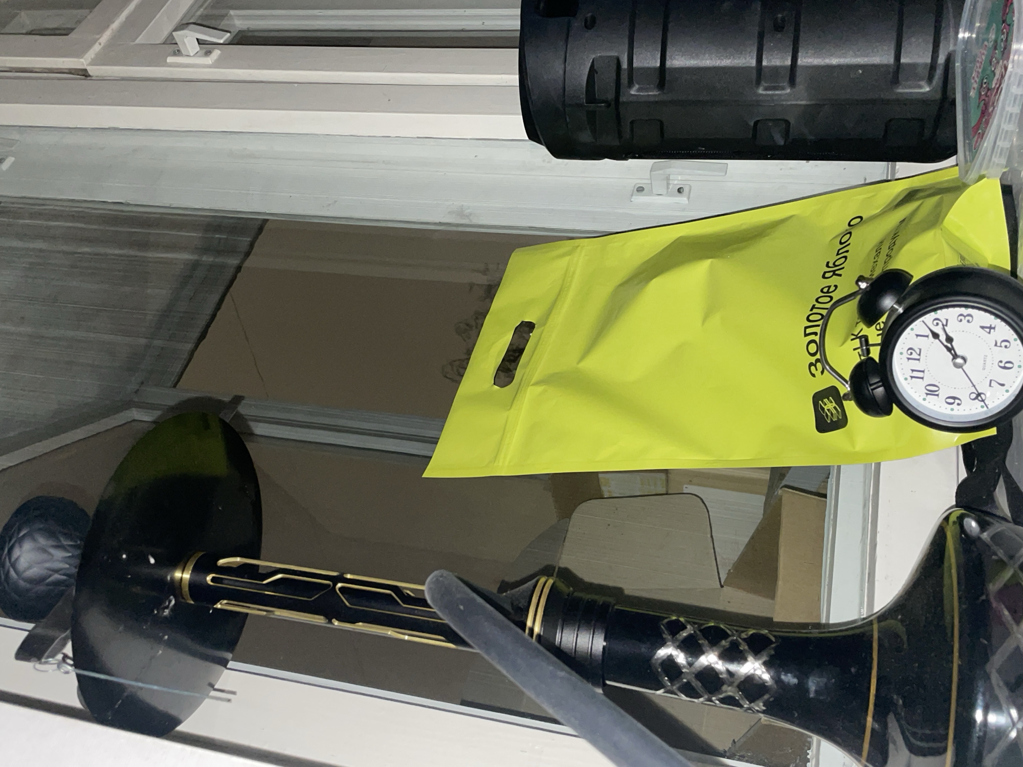 A still life image shows a hookah lying on a surface in front of a window, partially obscured by a bright yellow "Золотое Яблоко" shopping bag and a black alarm clock. The text on the bag is in Russian and translates to "Golden Apple." A black cylindrical object is visible in the upper right corner. The window frame is white, and the surface beneath the objects is a light neutral tone. The location, Murino, Russia is derived from the shopping bag.