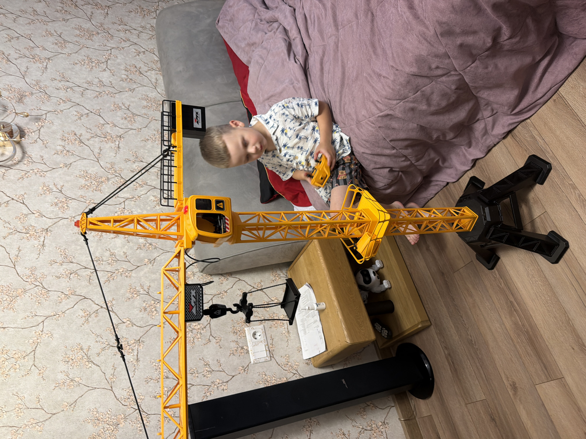 The image depicts a young boy playing with a large yellow toy construction crane. He is wearing a short-sleeved shirt with a palm tree print and patterned shorts. The boy is seated on the floor, near a large bed with a pink duvet. The crane is a prominent object in the scene, with its tall yellow lattice structure and black cables extending upwards. A black base with legs supports the crane.
On a wooden table next to the boy, there is a small white dog toy and some papers. The floor is made of wooden planks. The background shows a wall with floral wallpaper, a portion of a grey sofa, and a decorative lamp. The lighting suggests it is daytime. The overall scene appears to be an indoor setting, possibly a child's bedroom or play area in Minsk, Belarus. The boy seems engaged in his play with the toy crane. There is a black rectangular object with "CAUTION" written on it visible on the crane.