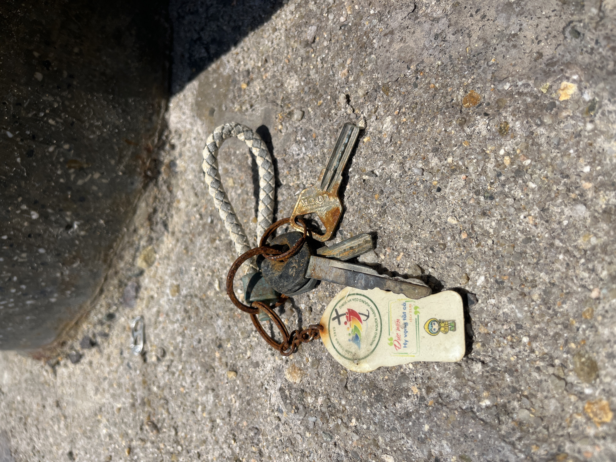 A set of heavily rusted keys, attached to a white, rectangular keychain via a rusty metal chain and rings, lies on a coarse, light gray concrete surface in Bac Nha Trang Ward, Vietnam. The keychain displays "NAM 2025", a graphic of a cross with three colorful figures, partially visible text "NHỮNG NGƯỜI HÀNH HƯƠNG", and further down, "Đức mến", "Hy vọng tất cả", "(1 Cr 13,8)", and a cartoon figure wearing a yellow raincoat and hat. One key is notably larger and dark with rust, while two smaller keys, one visibly marked "LOCK", are also severely corroded. A light-colored, braided fabric loop is attached to the main key ring. In the upper right background, a dark, textured, semi-cylindrical object, possibly a concrete pillar or base, casts a shadow across the lower right portion of the concrete ground. A blurred, shiny, metallic pull-tab object is visible in the upper left background. The scene is illuminated by direct sunlight, casting distinct shadows.