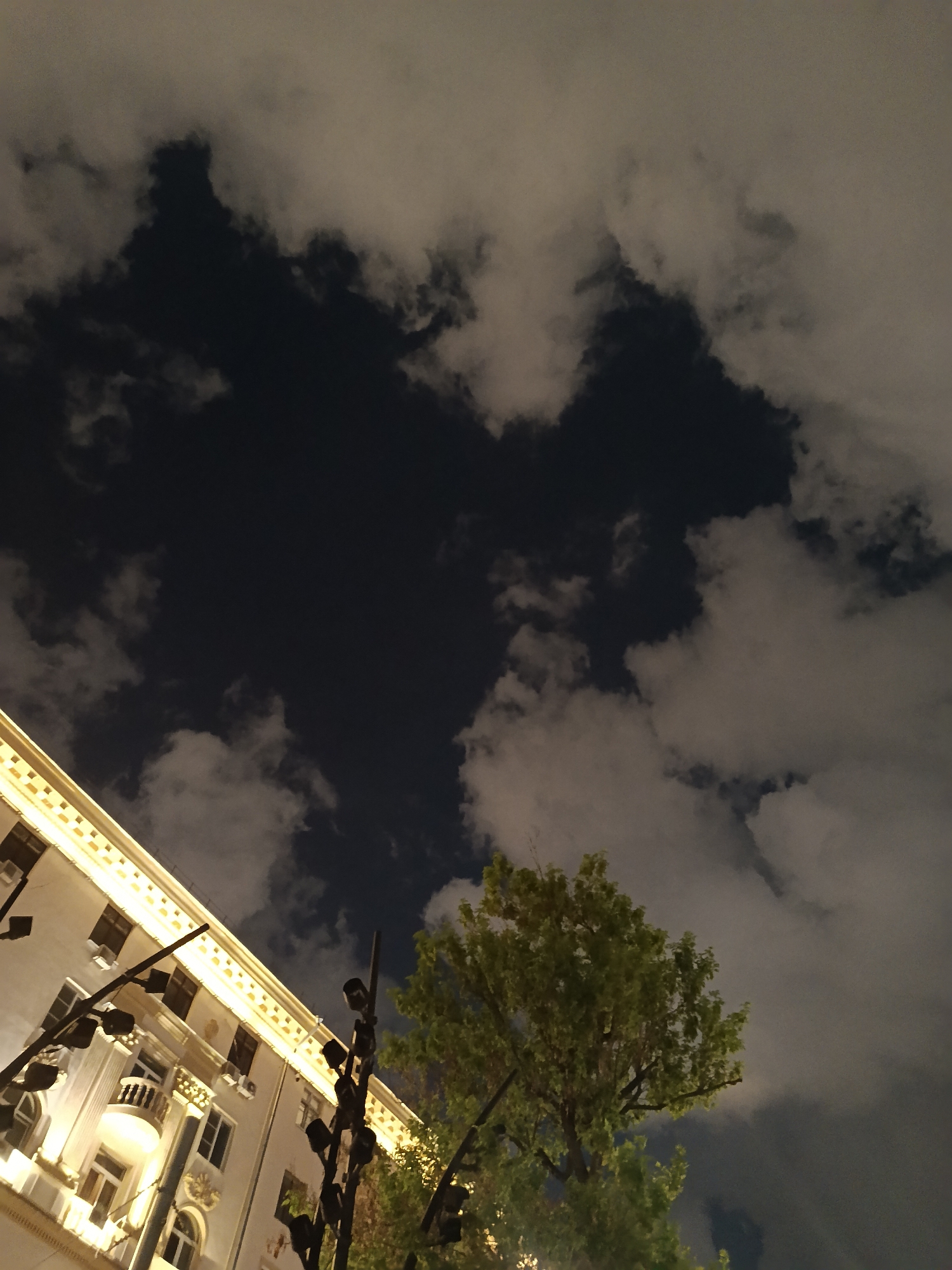 A low-angle shot depicts an urban scene at night, likely in Moscow, Russia. The sky is filled with dark clouds interspersed with lighter ones that may be reflecting light. A portion of a light-colored building with ornate detailing and illuminated windows runs along the left side of the frame. Streetlights are visible in front of the building, and a tree with green foliage is located near the right side of the frame. The overall impression is that the scene is dimly lit, with the building and clouds providing the primary sources of illumination.