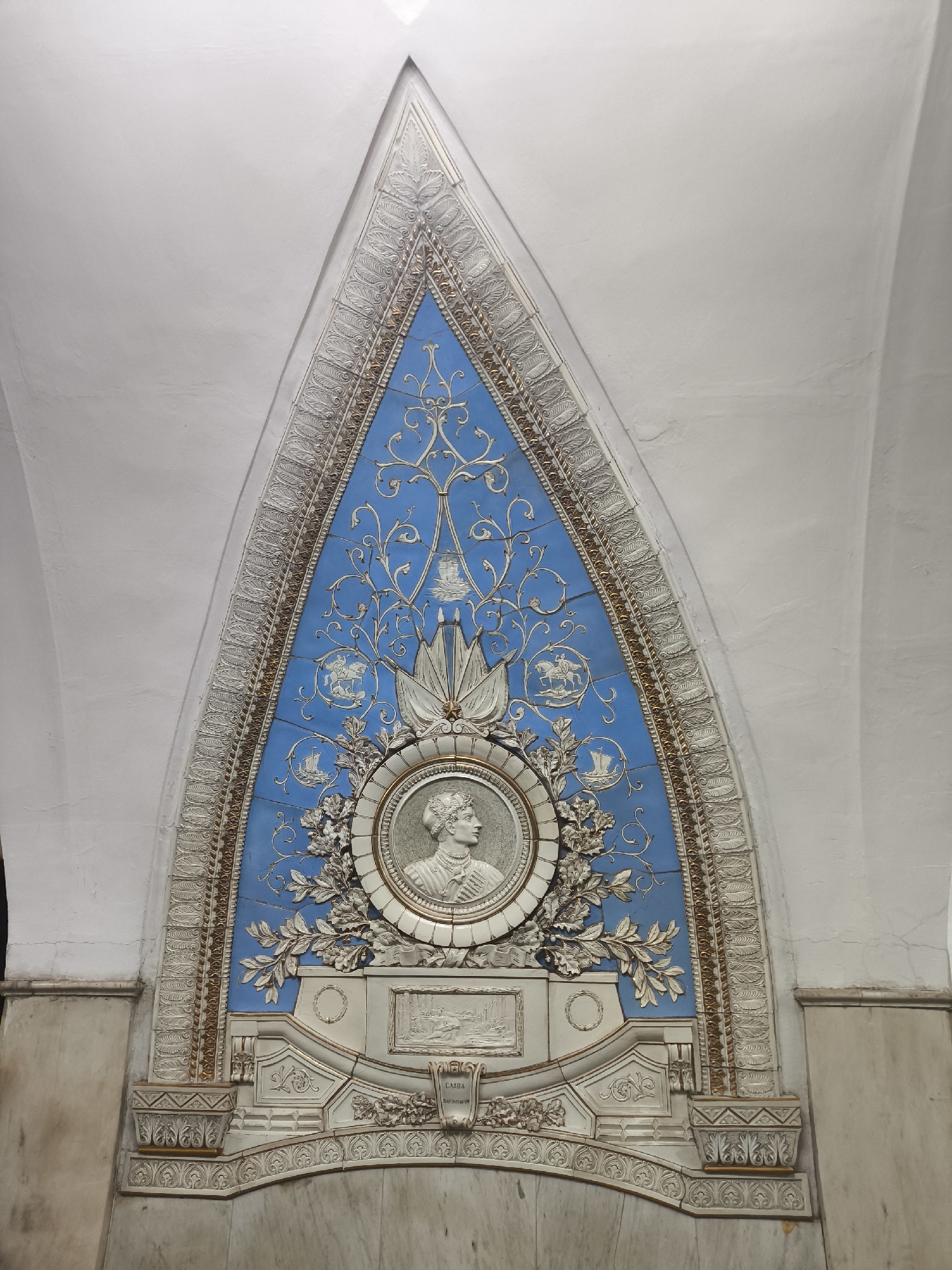 The image shows a decorative panel in an Unknown city, Unknown, likely within a subway or similar public space. The panel is predominantly a pointed arch shape and is inlaid into a white, curved wall. The central feature is a circular portrait in profile, possibly of a historical figure, surrounded by laurel leaves. This portrait is set against a light blue background decorated with swirling vine-like patterns and small emblems or symbols. A decorative border frames the blue section with a repeated leaf motif. Beneath the portrait, there's a structure resembling a base or pedestal with further decorative elements, including carved patterns and a small rectangular plaque. The material appears to be ceramic or a similar molded substance, with accents of gold color highlighting certain features. The wall itself is off-white and slightly textured.