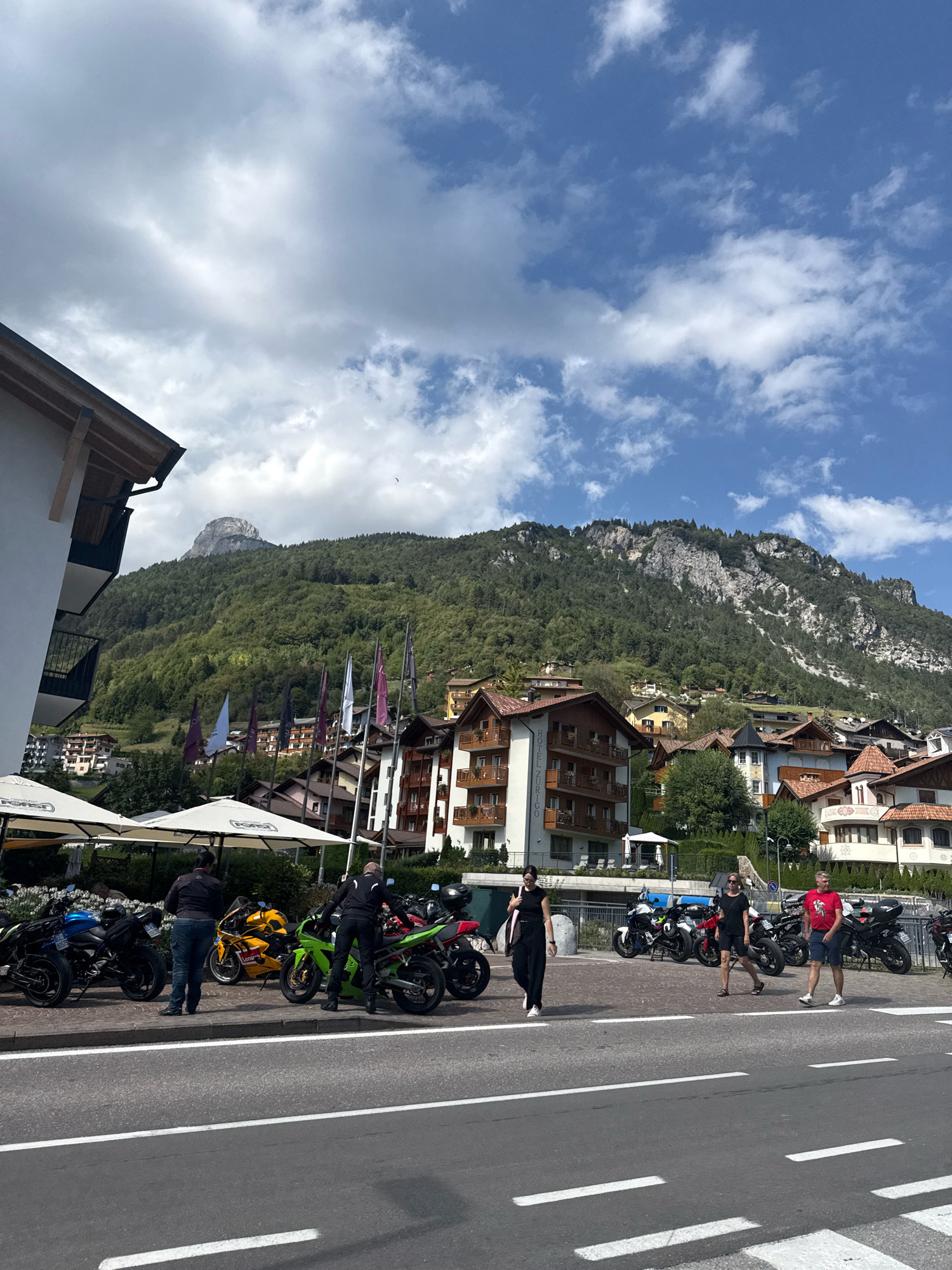 In the city of Molveno, Italy, a sunny day features a street scene with several parked motorcycles. In the foreground, a gray asphalt road has white lane markings. To the right of the road, a sidewalk with a curb is visible. On the sidewalk, five motorcycles are parked: a blue sportbike, a yellow sportbike, a green sportbike, a red sportbike, and a black motorcycle. Two people stand near the green motorcycle, and two other individuals walk along the sidewalk. Behind the sidewalk, white umbrellas are set up over tables, suggesting an outdoor dining area. Several flags are planted in the ground behind the umbrellas. In the background, multi-story buildings with traditional architecture are visible. A steep, tree-covered mountain rises behind the buildings, and a prominent, rocky peak is at the summit. The sky is blue with scattered white clouds.