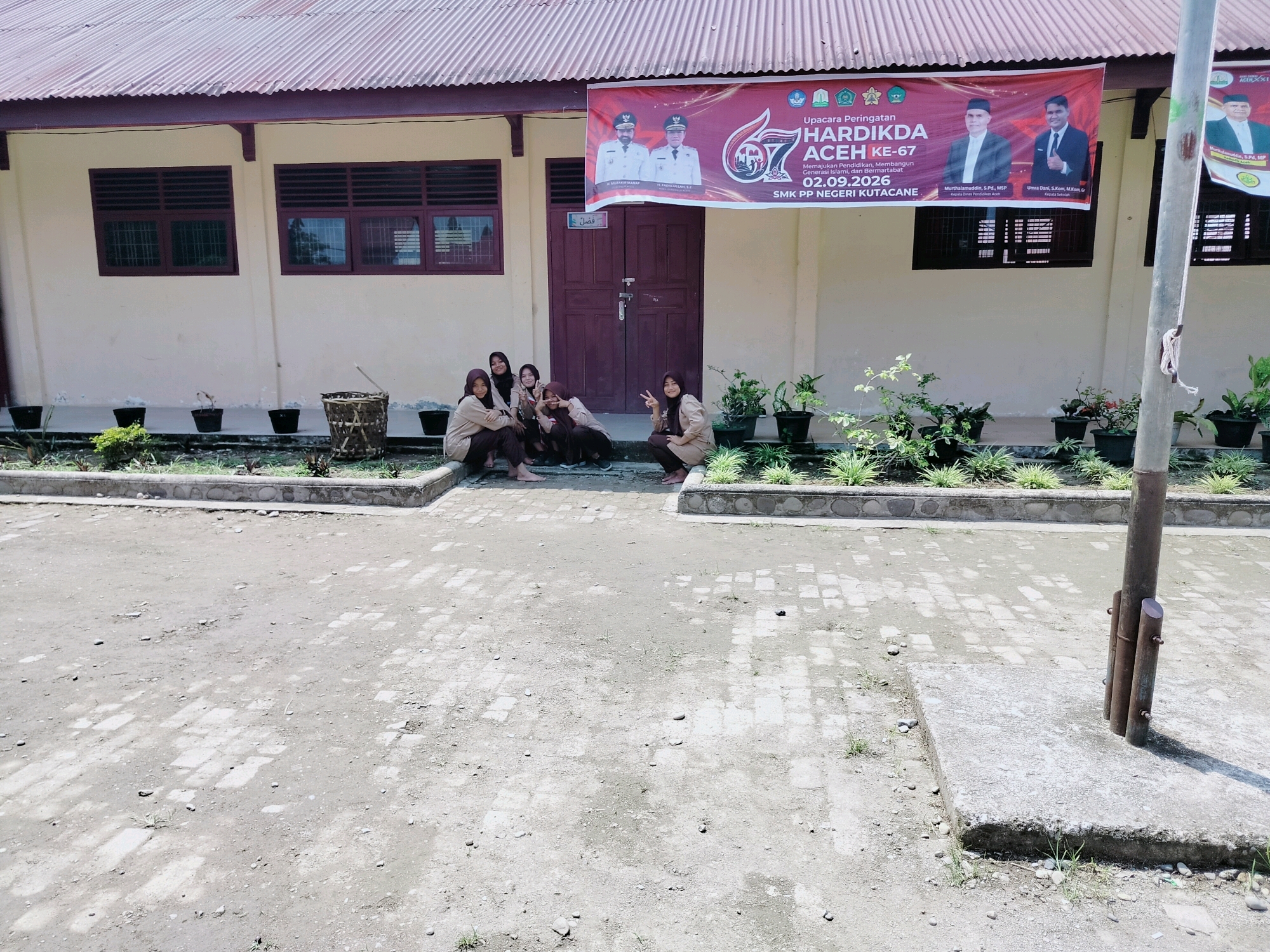 The image shows the exterior of a school building with a red, corrugated metal roof and cream-colored walls. A banner hangs across the front of the building, above a set of double doors. The banner features images of two men in suits, a logo, and text that appears to be in Indonesian, indicating a school event. In front of the building, several young women in school uniforms are gathered, some sitting and some standing, interacting and posing for the camera. The ground in front of them is a dirt and paved area with some scattered stones and vegetation. Several potted plants are visible along the wall of the building and in planters at the front. The overall scene suggests a casual gathering or event at an educational institution. The location is identified as Purwodadi, Indonesia, however, there are no visual cues in the image to confirm this specific location.