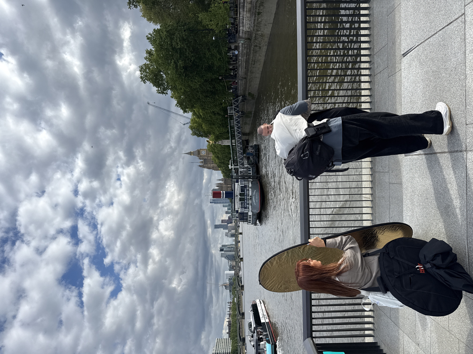 The image depicts a scene alongside the River Thames in London, United Kingdom. Two people are positioned near a black metal railing, with the river and city skyline visible in the background. One person, dressed in black pants and a white shirt, stands with a backpack and looks towards the river. The other person, holding a gold reflector, stands beside a large black bag. The Houses of Parliament and Big Ben are visible in the distance. Several boats navigate the river, and the sky is filled with numerous clouds.