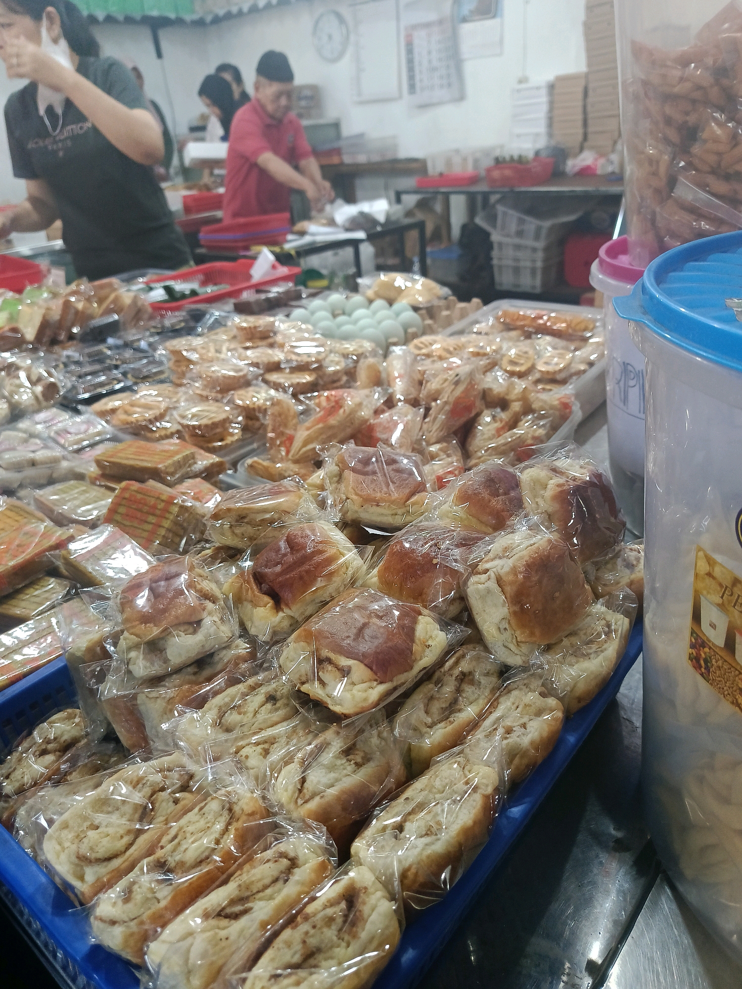 The image depicts a market stall in Yogyakarta, Indonesia, filled with various food items, primarily baked goods and snacks. In the foreground, a blue basket is overflowing with individually wrapped sweet rolls, possibly cinnamon rolls or similar pastries. Behind this, a variety of other packaged food items are displayed, including what appear to be layered cakes and other savory snacks. In the background, several people are visible, including a woman wearing a black t-shirt and a mask, and a man in a red shirt working behind a counter. The environment appears to be a busy food market or a small shop, with shelves and containers of food stacked in the background. The overall scene suggests a retail environment where food is being prepared or sold.