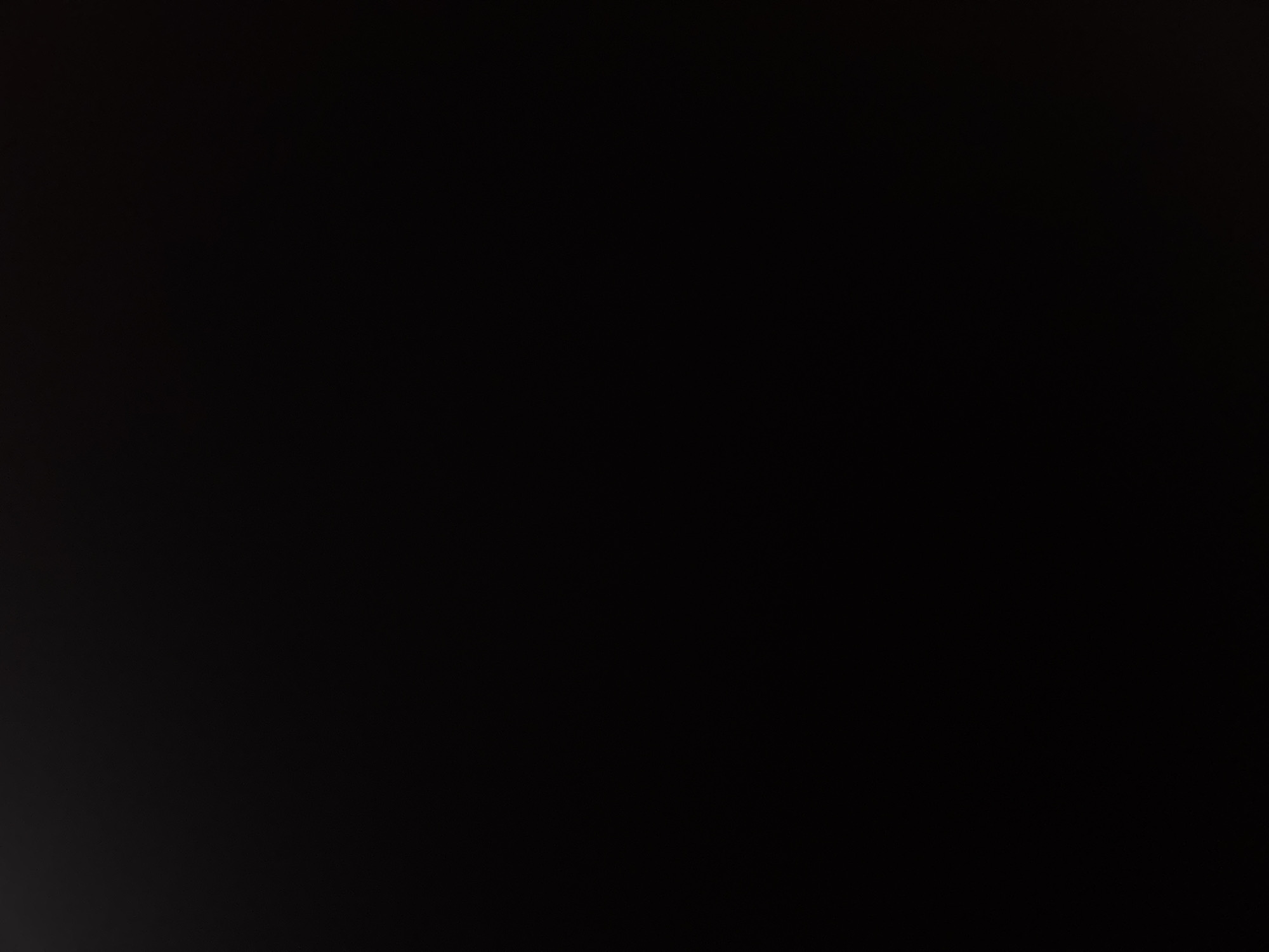 The image is severely underexposed, appearing almost entirely black with minimal discernible detail. Faint, dark grey and subtle reddish-brown hues are visible, primarily in the lower half of the frame. These hues reveal indistinct, elongated forms consistent with highly folded or draped fabric, possibly with a coarse texture, extending from the bottom edges towards the center. The upper portion of the image is pure black. No distinct objects, human figures, or specific actions are visible. The environment appears to be in complete darkness or captured with insufficient light. The described location of the scene is in Sofia, Bulgaria.