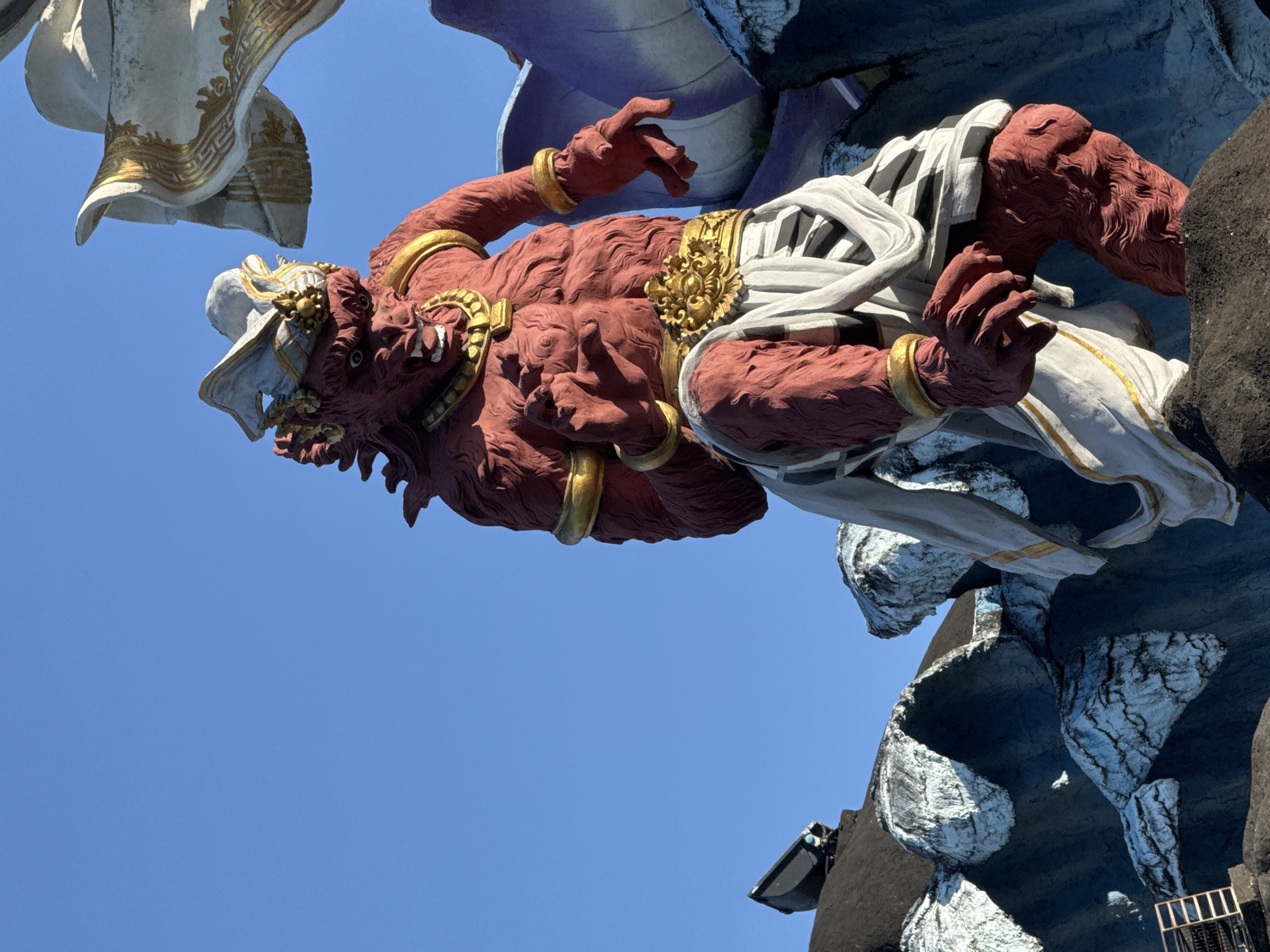 The image captures a portion of a large, ornate statue against a clear blue sky. The statue features a reddish-brown, muscular figure with golden armbands, a belt, and other golden ornamentation. The figure has a fierce, animalistic face with visible teeth and a decorated headdress. It appears to be wearing or partially draped in white fabric. The statue is set against a dark gray and white textured background, possibly representing rock or other architectural elements.
Given the style and location, it is likely that this statue is part of the Garuda Wisnu Kencana Cultural Park in Denpasar, Indonesia.