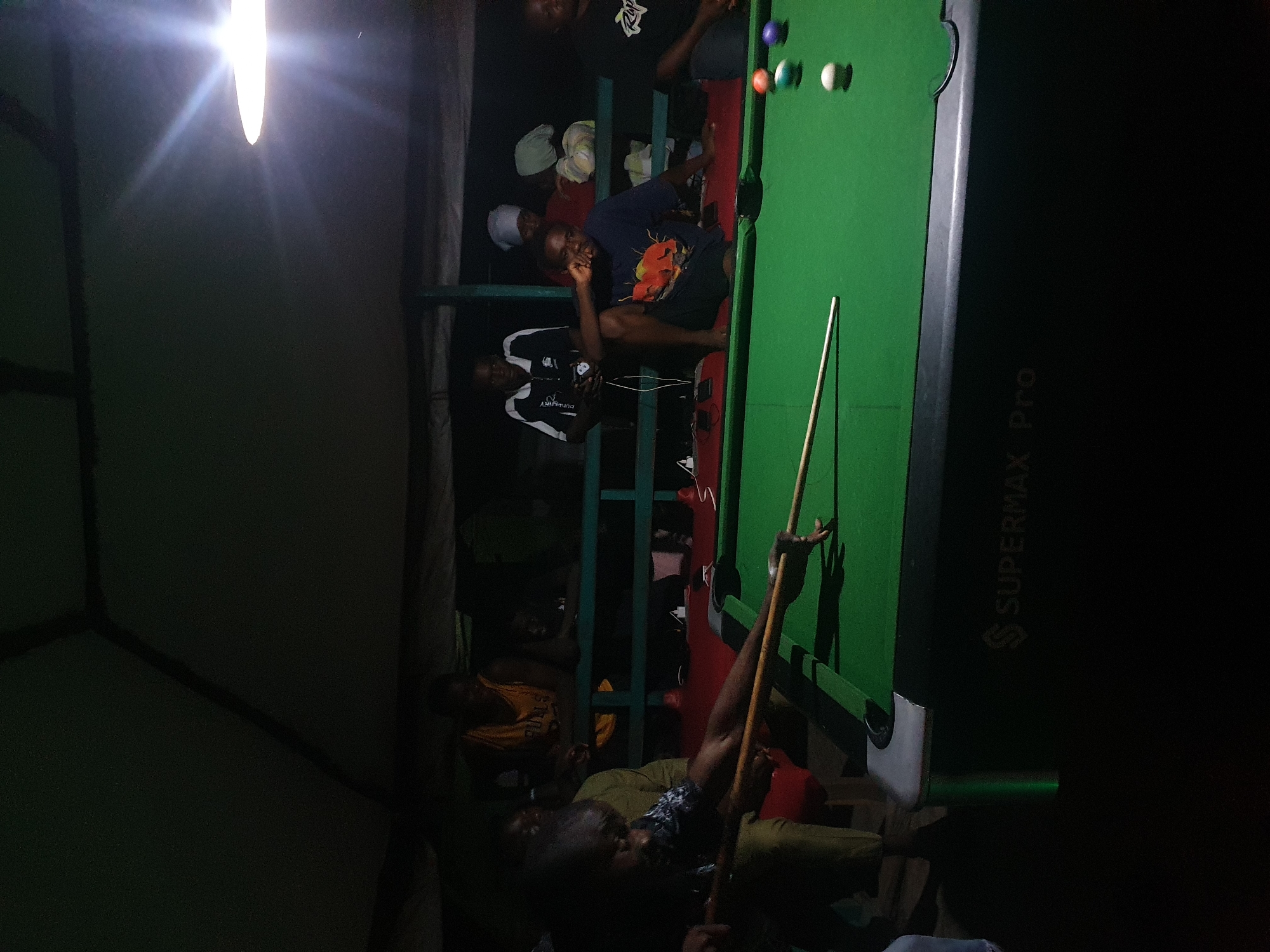 A group of individuals are gathered around a green pool table in what appears to be an outdoor, covered area at night. The lighting is dim, with a bright overhead light illuminating the pool table and a portion of the surrounding space. Several people are visible, some standing and others seated.
In the foreground, a person is actively playing pool, holding a cue stick and aiming at a ball on the table. The pool table has several colored balls scattered across its surface. Behind the player, other individuals are observing or waiting their turn. One person in a yellow basketball jersey is visible, leaning on a railing. Other figures are seated on benches or the edge of the pool table. The environment suggests a recreational setting, possibly a bar or a communal gathering spot. The pool table itself is marked with the brand "SUPERMAX Pro". The overall atmosphere is casual and social. The image was taken in Lokoja, Nigeria.