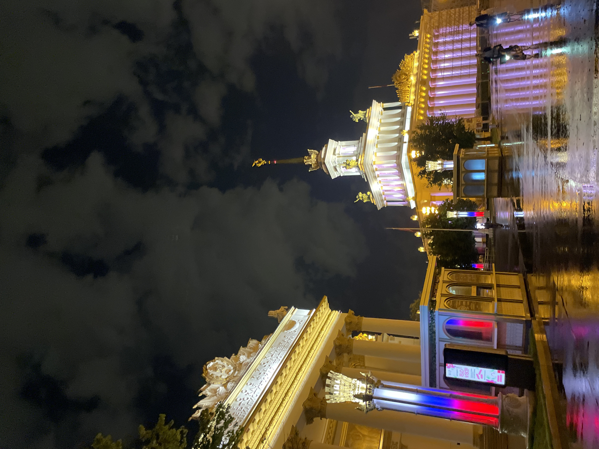 This nighttime photograph captures a grand architectural scene in Moscow, Russia, showcasing illuminated buildings against a dark, cloudy sky. The foreground features ornate structures with bright, yellowish lighting that highlights their classical design, including prominent columns.
A central, white and gold structure reaches upward, adorned with statues and bathed in soft purple lighting. Another building behind it is illuminated with violet lighting. The ground appears wet, possibly due to rain, with reflections of the bright lights enhancing the scene.
The upper portion of the image is dominated by a dark, cloud-filled sky. Distant figures can be made out, suggesting the presence of people in the vicinity. The scene conveys a sense of grandeur and architectural beauty in Moscow.