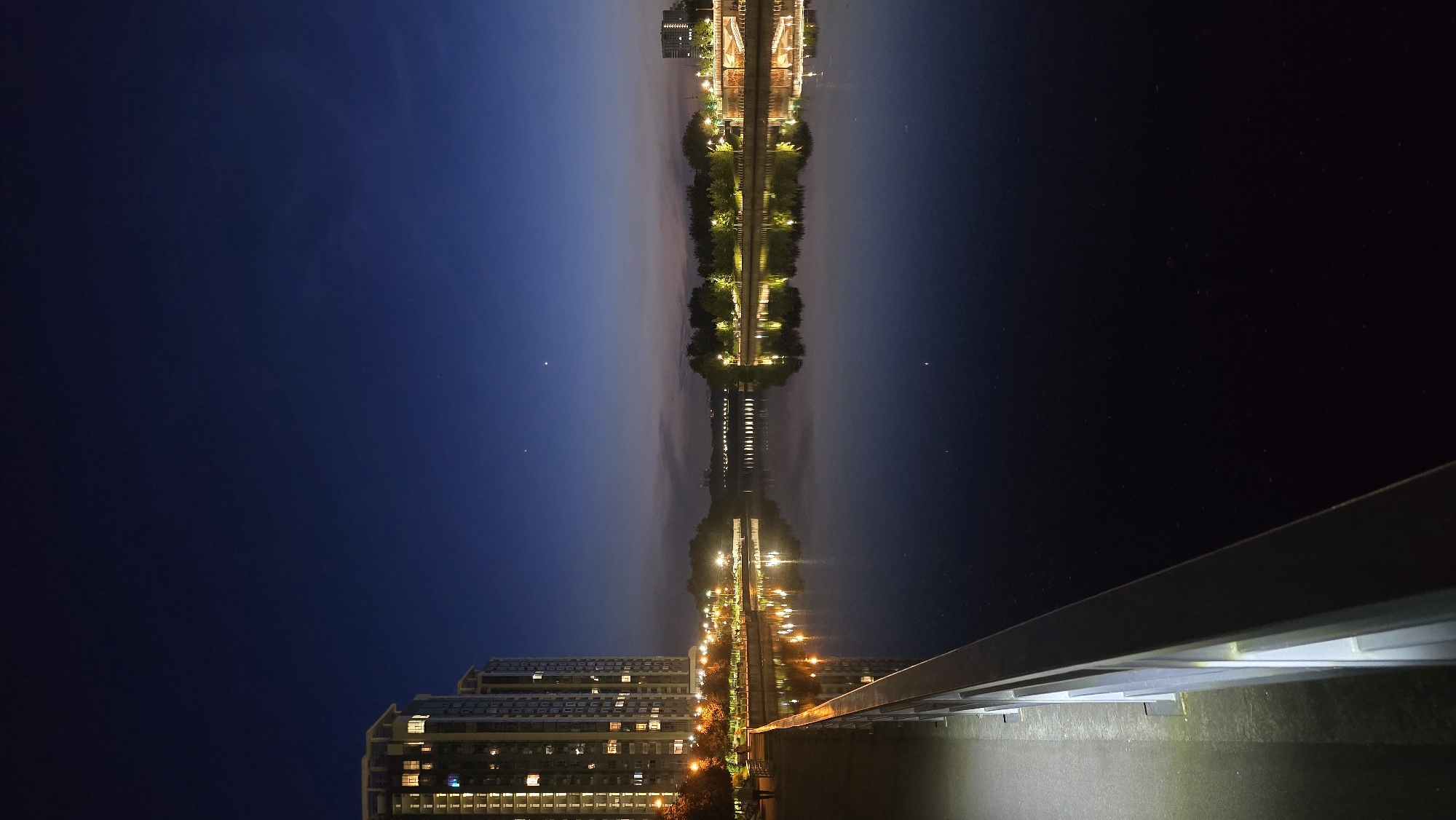 The image, when viewed in its correct landscape orientation, captures a serene nighttime or deep twilight urban scene in Kyiv, Ukraine, dominated by reflections on a body of water.
The central feature is a long, narrow stretch of land or an elevated structure, possibly a bridge or embankment, adorned with numerous bright lights and trees. This illuminated strip runs horizontally across the middle of the frame, creating a radiant line of light that stands out against the dim surroundings. Below this structure, the calm water perfectly reflects the lights and elements above, creating a near-symmetrical mirrored image.
To the far left of the main illuminated strip, a multi-story modern building with many brightly lit windows is visible, suggesting an apartment complex or office building. Its lights are also clearly reflected in the water below. The sky above is a deep, dark blue, transitioning to black on the far right, with a few faint celestial objects or distant lights barely visible.
In the bottom right corner, a portion of a railing or barrier extends into the foreground, indicating the viewpoint is from another elevated structure, possibly a bridge or promenade overlooking the water. No people are visible, and no specific activity is taking place, lending the scene a quiet, contemplative atmosphere. There is no visible text in the image. The overall impression is one of urban tranquility and illuminated beauty at night.
