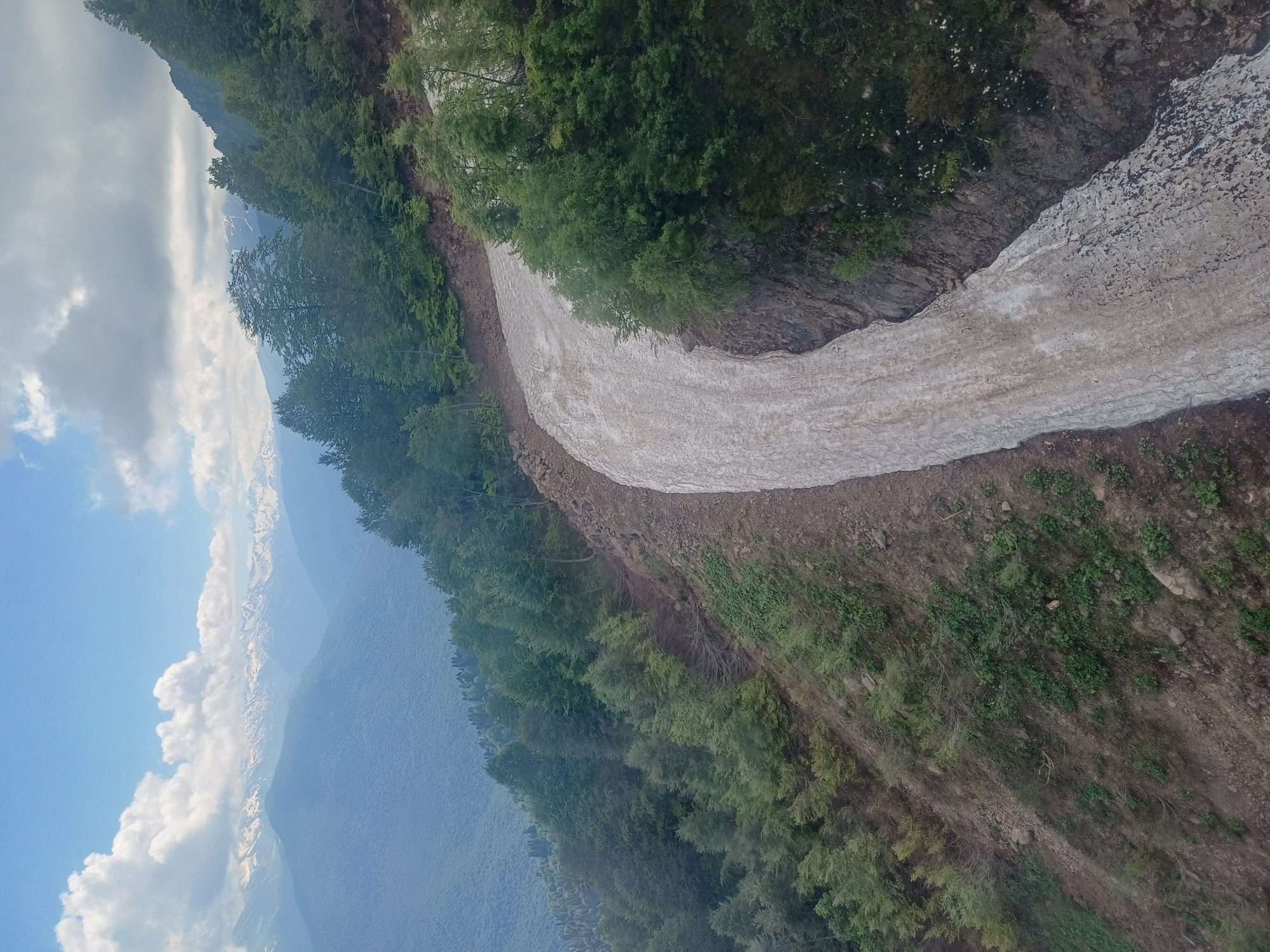 A high-angle, wide shot captures a winding path of dirty snow on a steep mountain slope. The snow is discolored with dirt and debris, indicating melting and a natural transition from winter to spring. Dense green trees cover the surrounding mountainside, their foliage vibrant against the pale snow. In the distance, a range of snow-capped mountains stretches across the horizon under a partly cloudy blue sky. The scene is devoid of any people or animals, presenting a serene and natural mountain landscape. The location is identified as the Esto-Sadok resort area in Russia, known for its mountainous terrain and ski slopes.