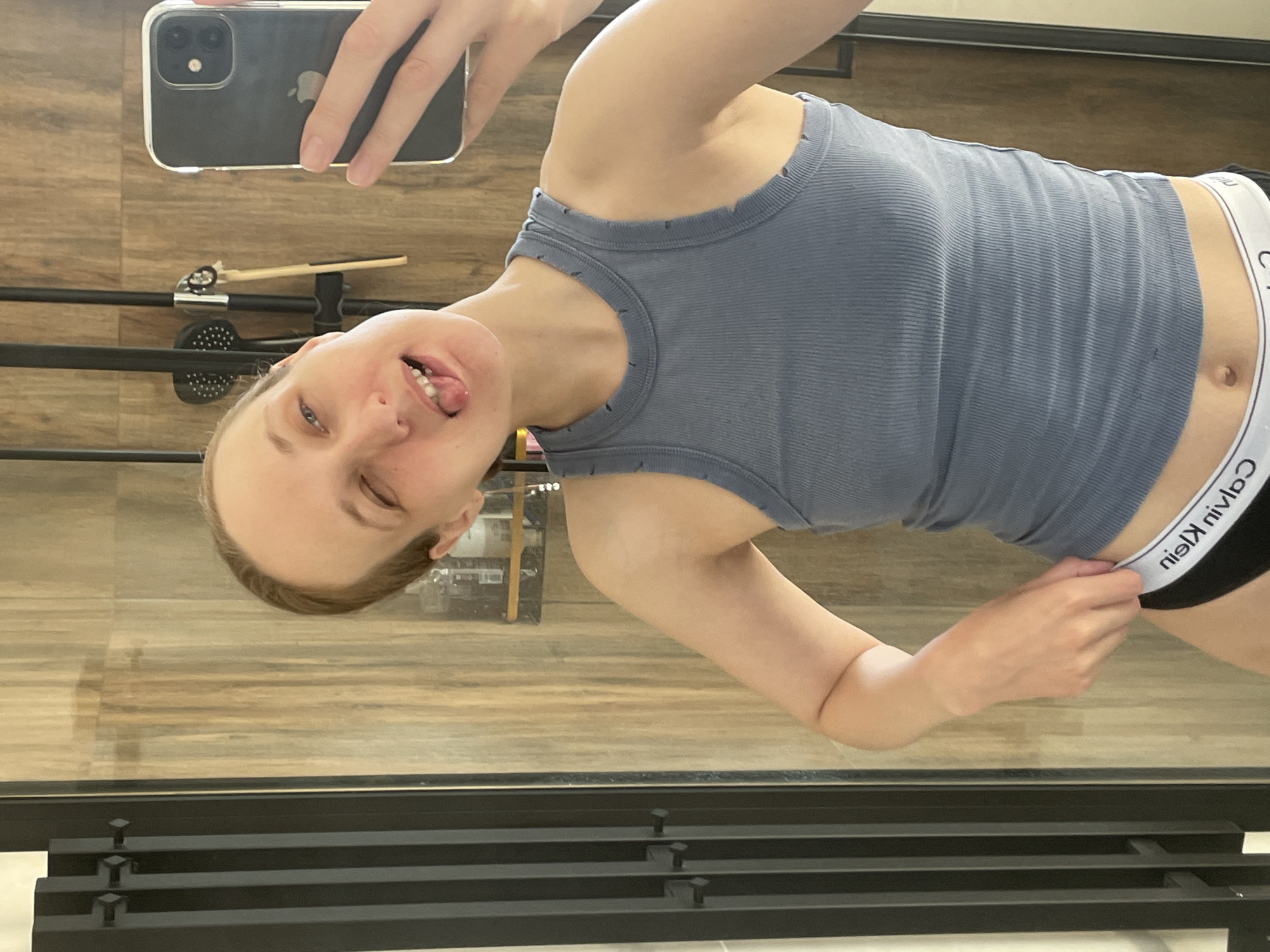 A fair-skinned person is holding a smartphone in their left hand, taking a selfie. The person is wearing a grey tank top and black underwear with a white Calvin Klein waistband. The person is sticking their tongue out. The background appears to be a room with wood-patterned flooring and a black metal structure with horizontal bars. There is no visual information to definitively locate the scene in Подшивалово, Russia.
