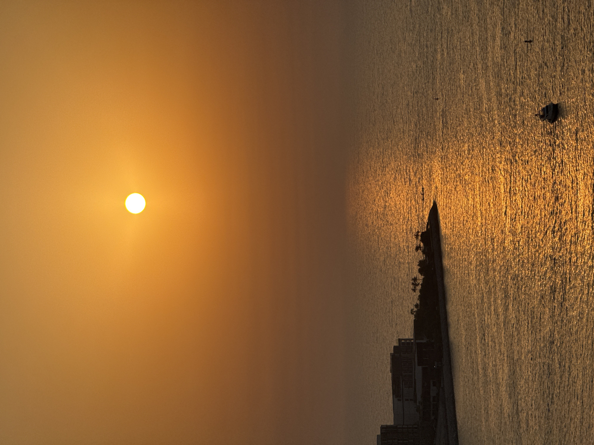 This vertical, eye-level, medium shot captures a sunset over the Arabian Gulf in Dubai, United Arab Emirates. The sun, a bright white orb, hangs in the upper center of the frame, casting a warm orange glow across the sky and the rippling water. The light creates a shimmering path on the sea, reflecting the sunset's hues.
In the lower left quadrant of the image, a modern architectural development lines the coastline. The buildings are angular and dark, silhouetted against the vibrant sky. Some palm trees are visible along the edge of the land. Further out in the water, a small motor yacht, dark against the bright reflections, sails peacefully. Two small buoys or markers are visible in the distance, one to the left of the yacht and another to its right. The overall atmosphere is serene and picturesque, characteristic of a coastal sunset in a metropolitan area.