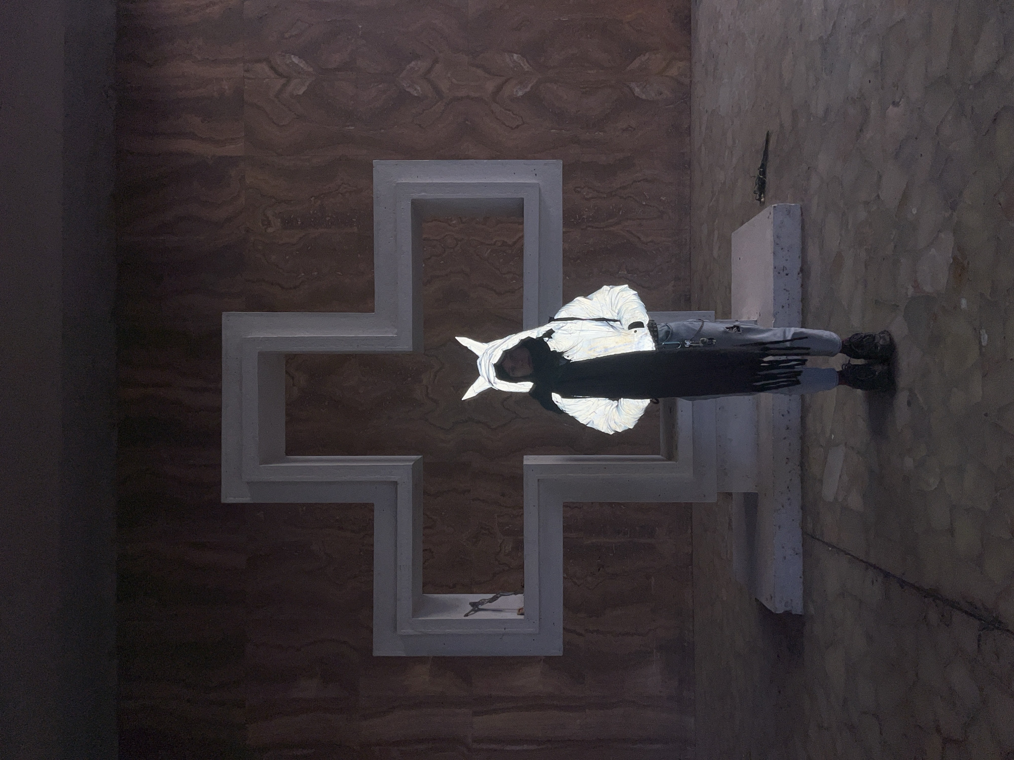 A person wearing a reflective jacket and dark pants lies across a large, white, stylized cross made of concrete. The cross is mounted on a patterned brown wall. A small object rests inside the lower arm of the cross. The person's lower body extends past the edge of the cross, towards a speckled stone floor.