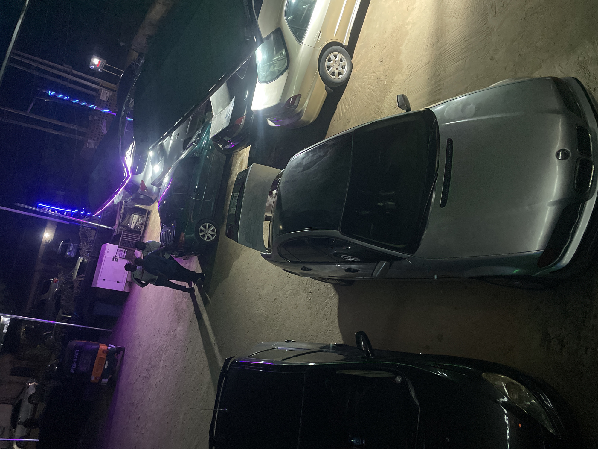 The image depicts a nighttime scene in Gura Top, Nigeria. Several cars are parked on a paved surface. The vehicles include a silver car with its hood open, a black car, a tan car, and a dark green car. Two people are walking between the cars and a white generator. In the background, there is a structure with blue and purple neon lights. There is a small, orange and black vehicle. The scene is generally dark, lit by artificial light sources.