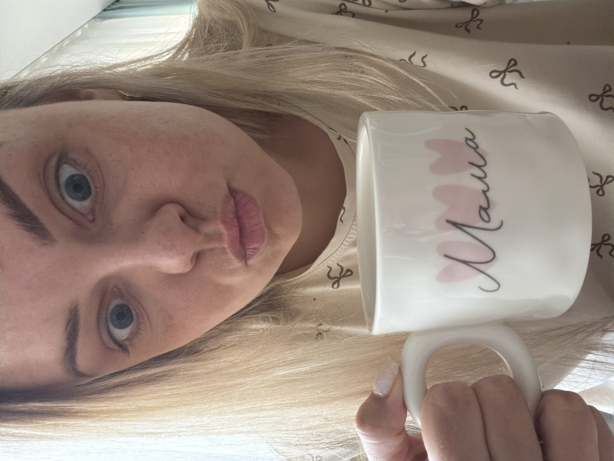A blonde woman with fair skin is holding a white mug with the word "Mama" written in Cyrillic script with pink hearts. Her face is turned to the left, and she is making a kissing face. She is wearing a light-colored shirt with a bow pattern. The background is out of focus, but suggests an indoor setting. The location in the city of Votkinsk, Russia, is not visually discernible from the image.