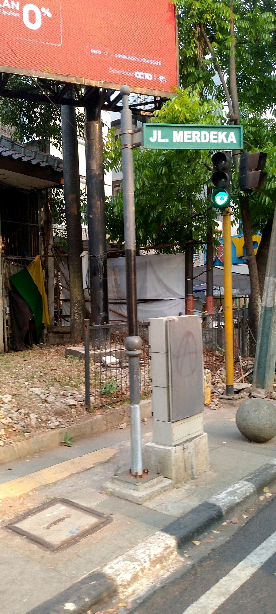 The image shows a street scene in Bandung City, Indonesia, featuring a street sign that reads "JL. MERDEKA". A traffic light is visible, currently showing a green light. In the background, there is a large red billboard with white text, partially obscured by trees. The sidewalk has a manhole cover and a concrete utility box with a spray-painted symbol. A large, round concrete ball is also present on the sidewalk. The environment appears to be urban, with trees and a building visible. The overall scene is of a typical city street.