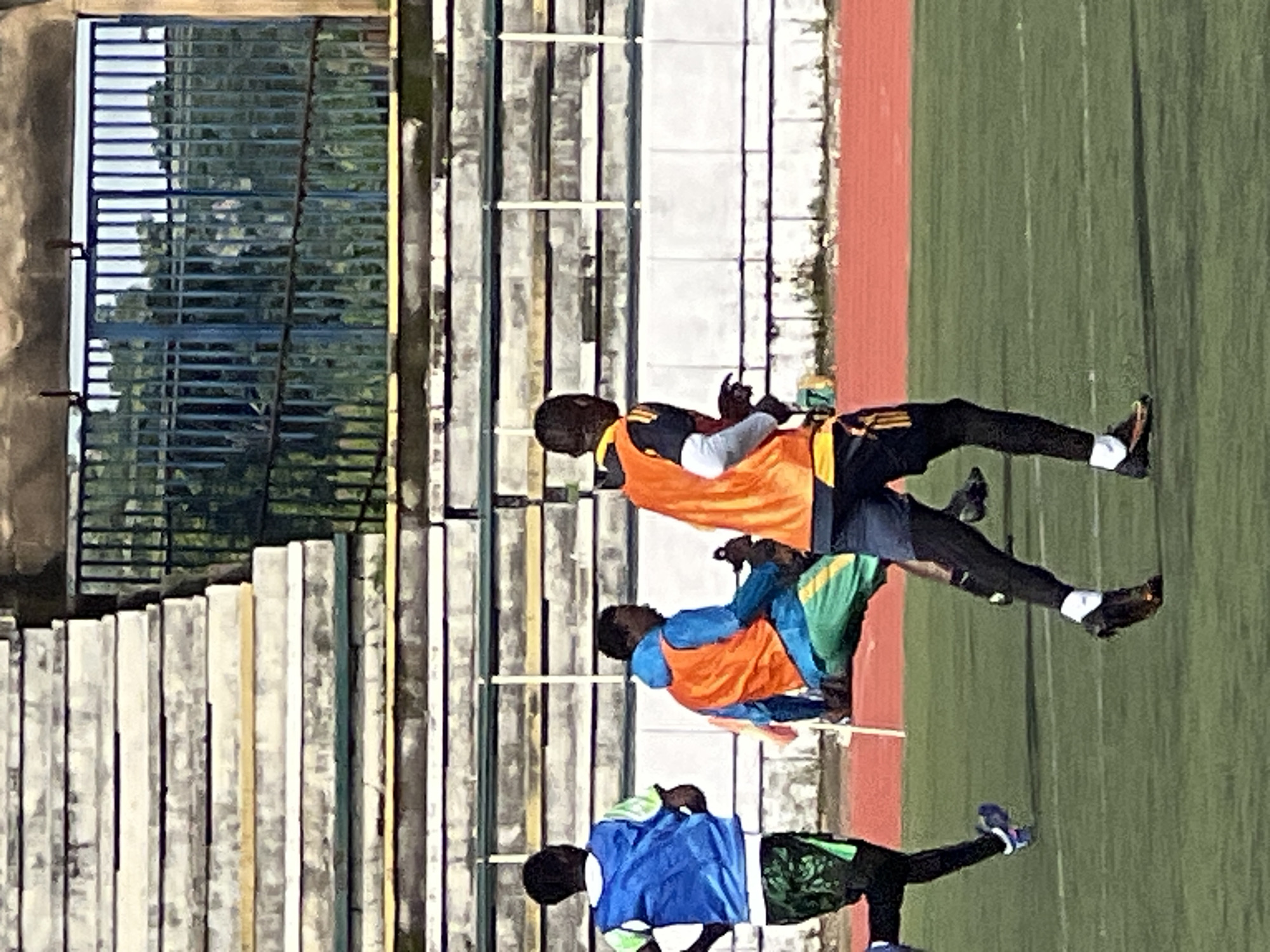 This image depicts three individuals on a green turf field, likely a sports ground, with stadium seating in the background. The stadium features layered concrete benches with yellow and green accents, and a blue metal fence behind them. The individuals are dressed in athletic wear. One person on the left wears a blue vest over a t-shirt and green patterned shorts. In the center, another person is crouched or sitting, wearing a blue hooded jacket with an orange vest and green pants. The person on the right is running forward, wearing an orange vest over a black shirt with yellow accents and black shorts. The field itself is marked with white lines, suggesting a sports field. The overall scene is likely taking place in New Stadium, Nigeria, given the context.