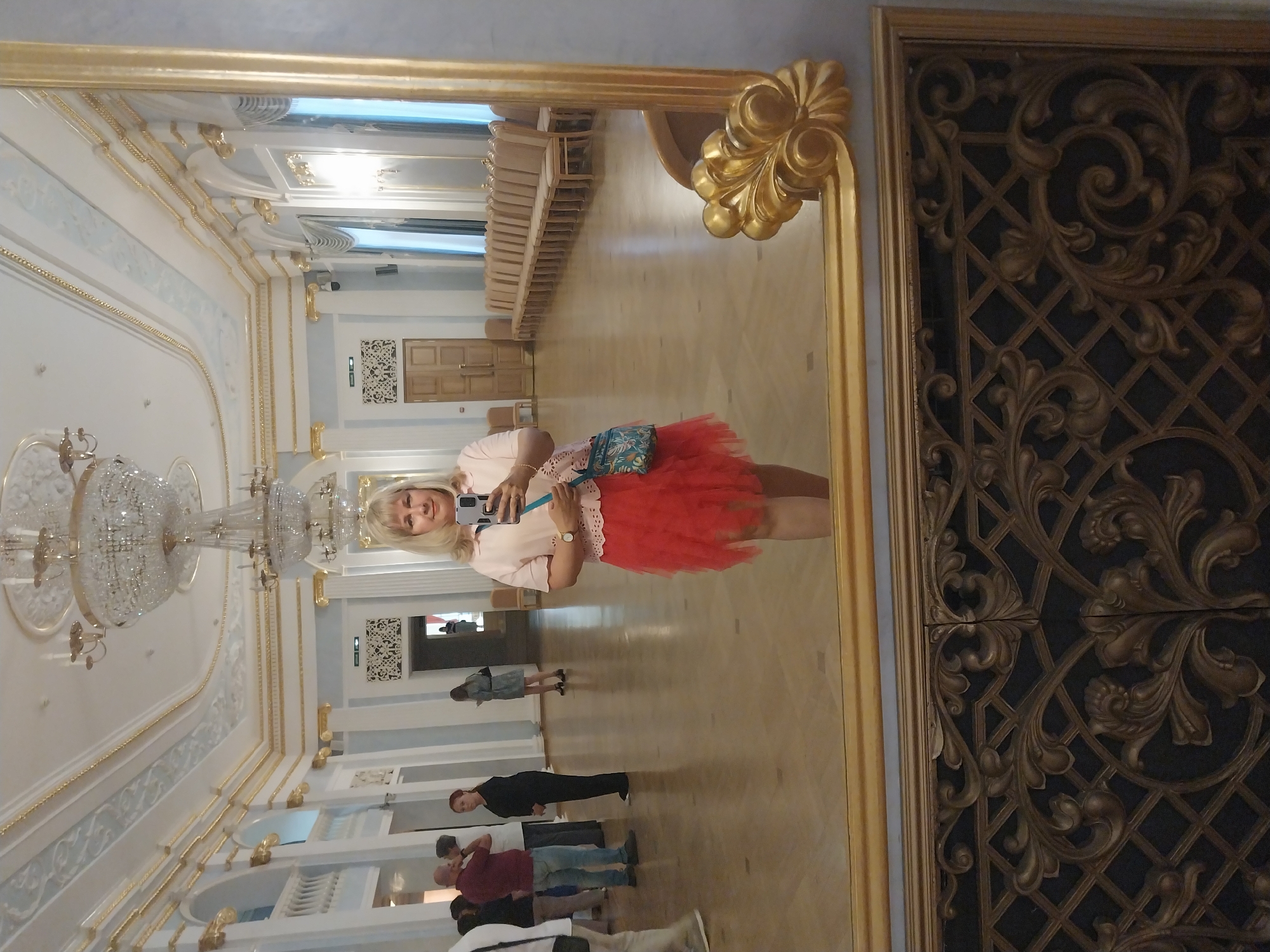 In Orenburg, Russia, a woman with blonde hair, wearing a light pink blouse and a red tulle skirt, stands in a grand, ornate hall, taking a selfie in a large, gilded mirror. The hall features high ceilings with elaborate chandeliers and decorative moldings, large windows with draped curtains, and a polished wooden floor. Several people are visible in the background, some appearing to be tourists or visitors. The woman is holding a smartphone and a small, patterned bag. The lower part of the image shows a detailed, decorative grille.