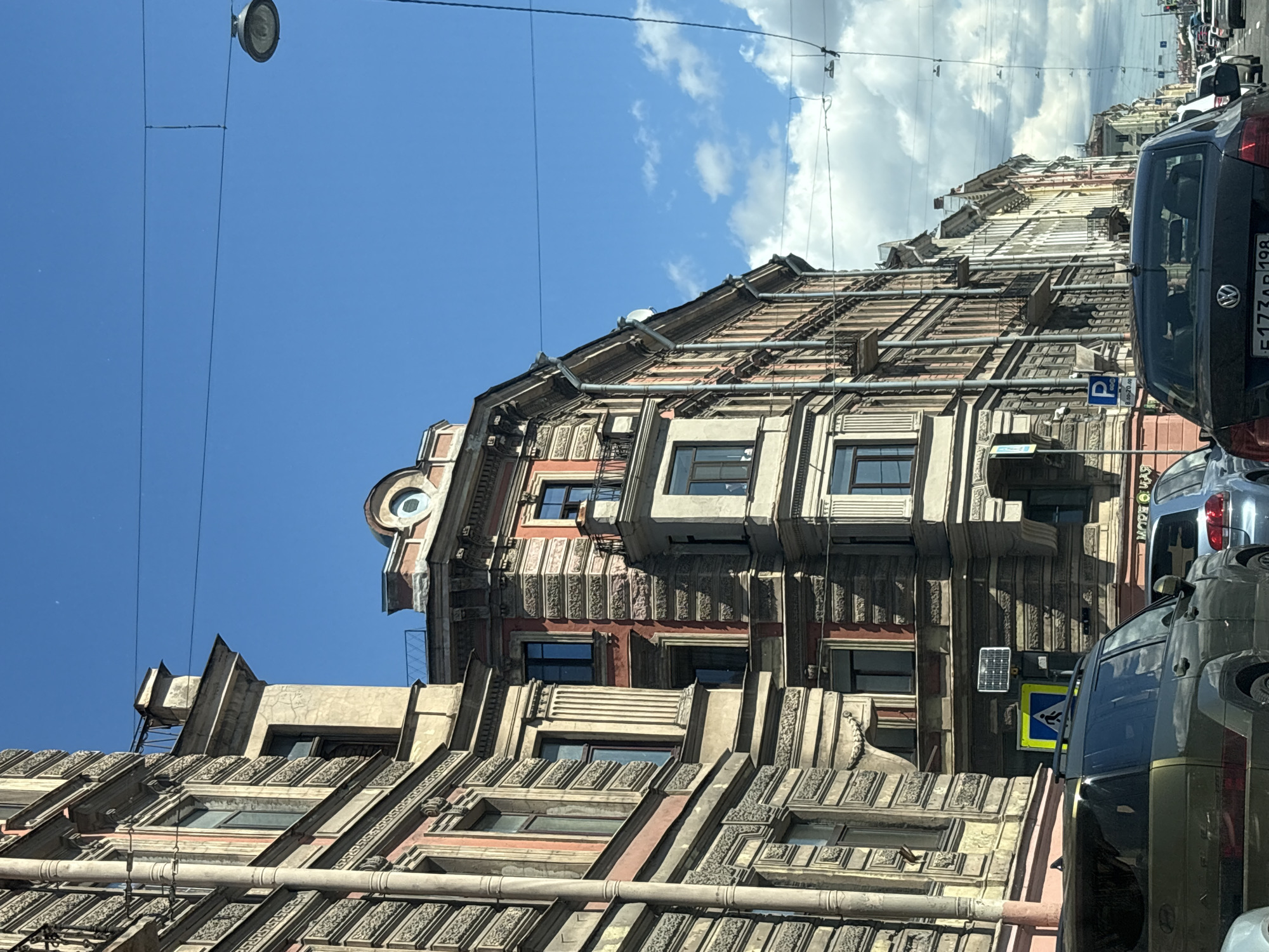 The image captures a street view in Saint Petersburg, Russia, dominated by the ornate architecture of historic buildings lining a road. The buildings exhibit classical architectural styles with detailed facades, balconies, and numerous windows. The sky is a bright blue with scattered white clouds. Power lines and a street lamp are visible overhead. Several cars are parked and in motion on the street, including a dark-colored SUV in the foreground and a dark sedan with a partial license plate visible. Traffic signs, including a pedestrian crossing sign and a parking sign, are present along the roadside. The overall scene suggests an urban environment with a blend of historical and modern elements.