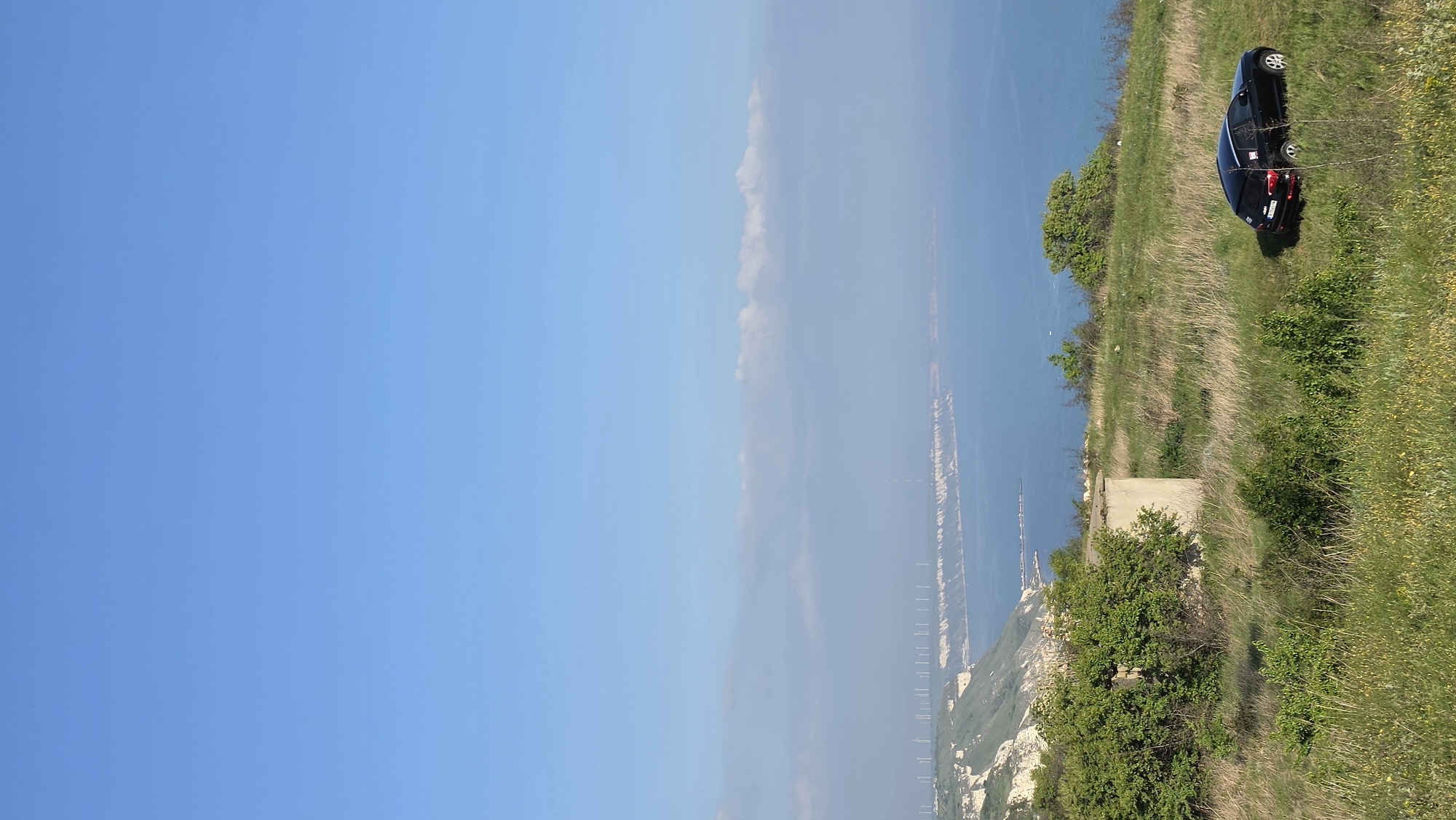 The image depicts a coastal scene in Bojurets, Bulgaria, featuring a black car parked on a grassy cliff overlooking the Black Sea. The sky is predominantly clear blue, with a faint line of clouds on the horizon. The coastline is visible with a pier extending into the sea. The foreground shows a mix of grass, bushes, and a concrete structure.